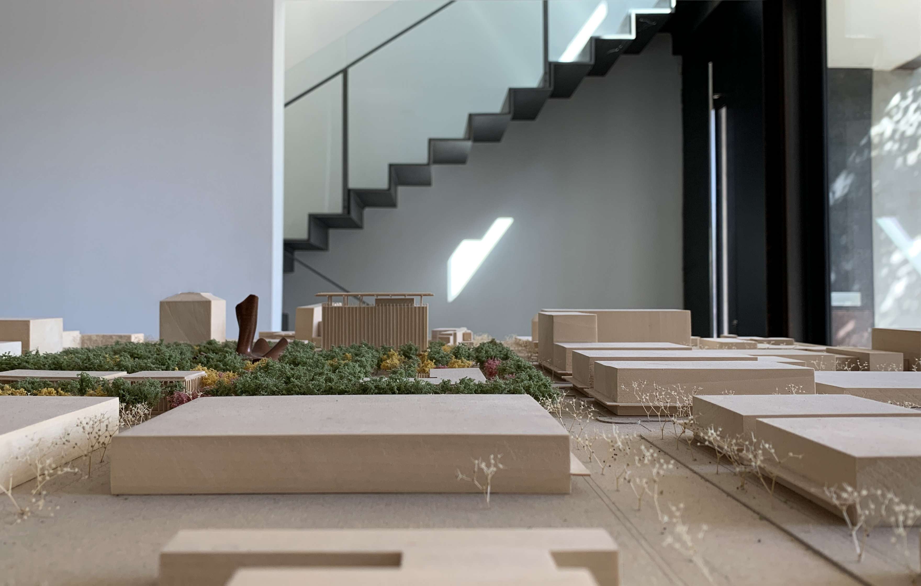 A close-up of a detailed architectural model with various small buildings and greenery, placed indoors near a modern black staircase and a large window.