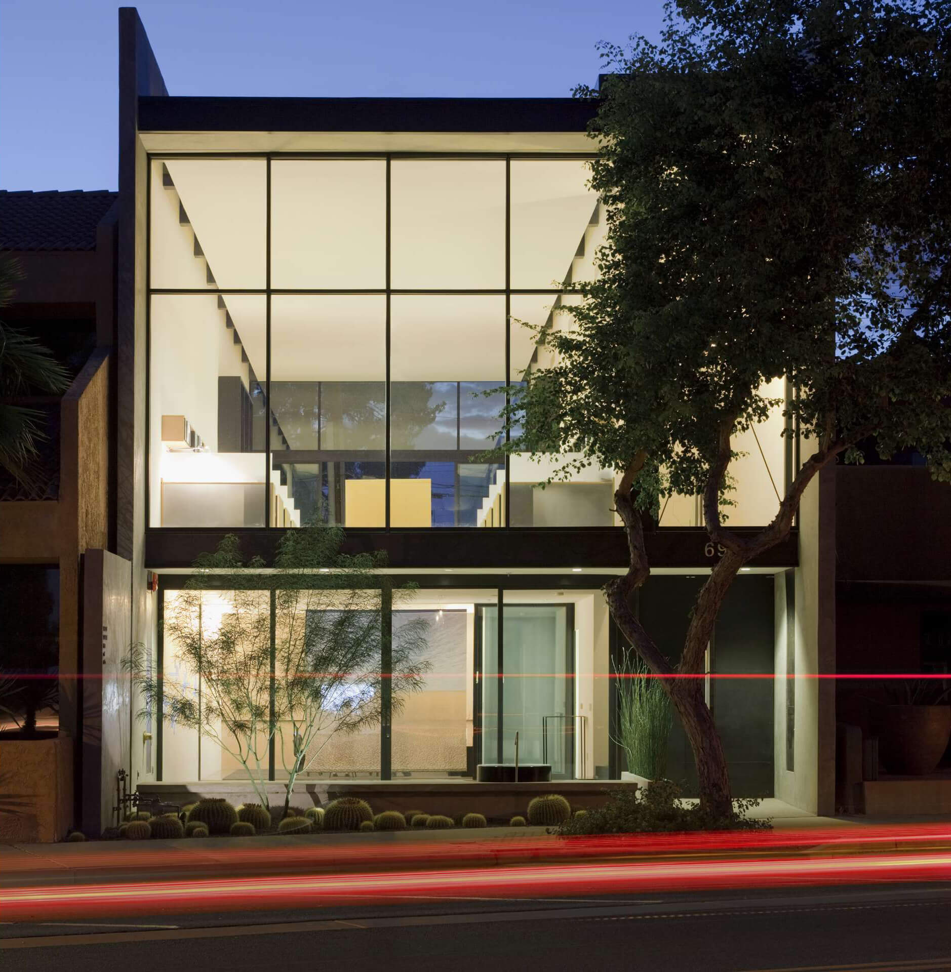 A modern two-story building with large glass windows, illuminated from the inside, with a tree in the front yard and a blurred light trail from passing traffic in the foreground.