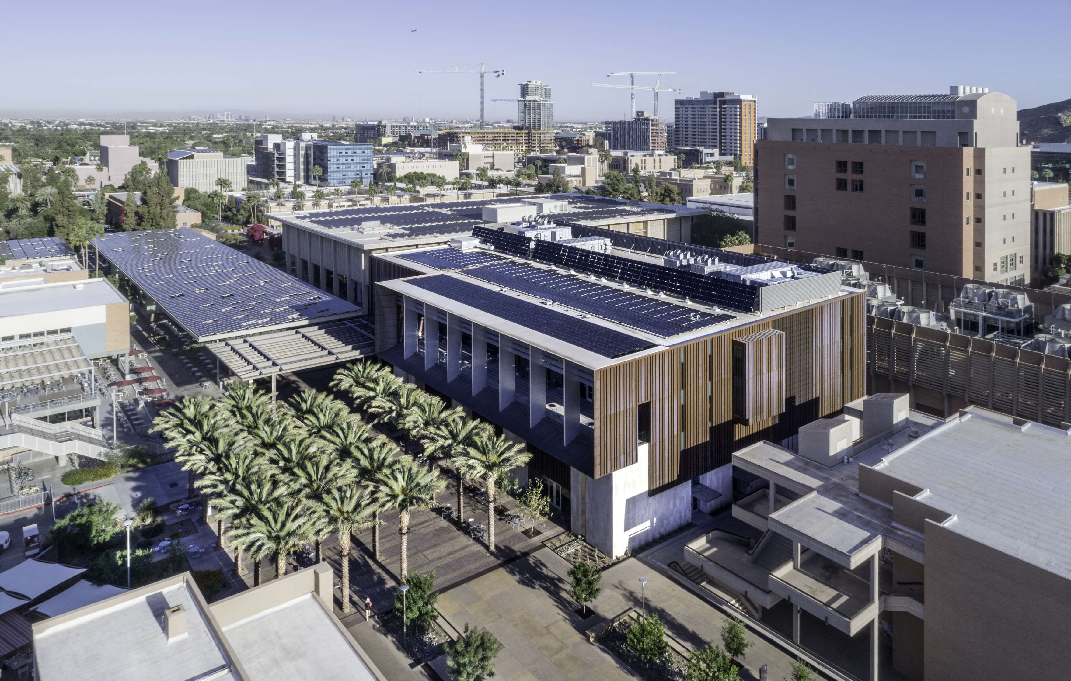 Aerial view of an urban area featuring a large, modern building with solar panels on the roof, surrounded by palm trees and neighboring structures. City skyline visible in the background.