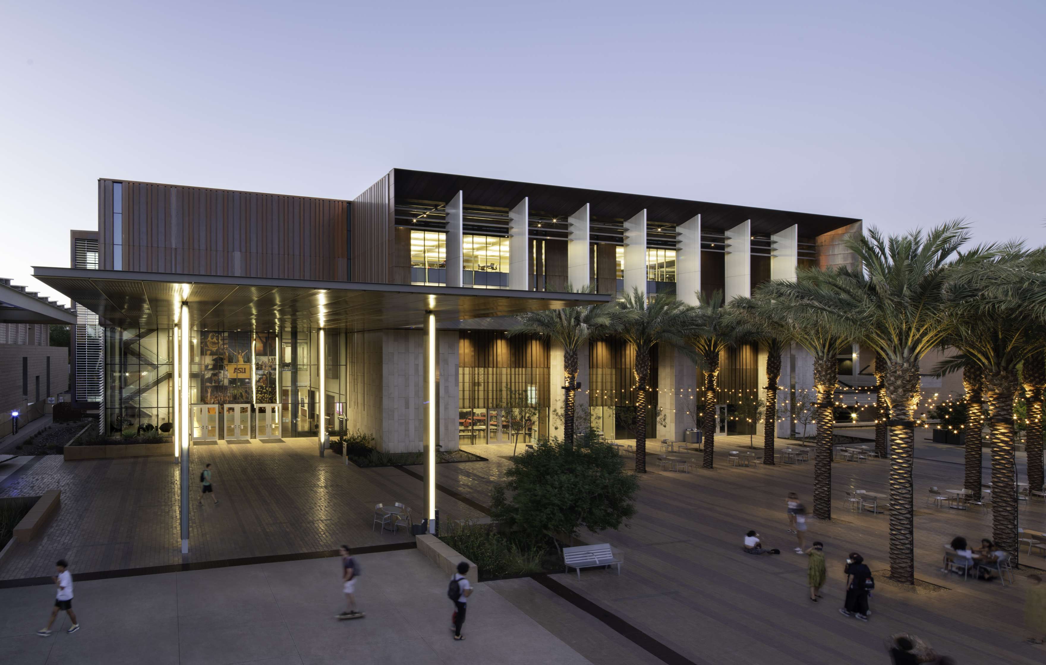 A modern building with large glass windows and a columned entrance, situated next to a plaza lined with palm trees adorned with string lights. The scene is captured at dusk with a few people walking.