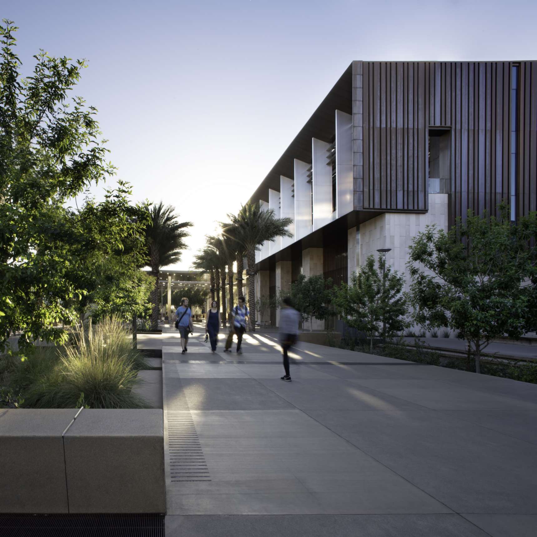 Several people walk along a pathway next to a modern building with large vertical panels and surrounded by trees and greenery, at sunset.