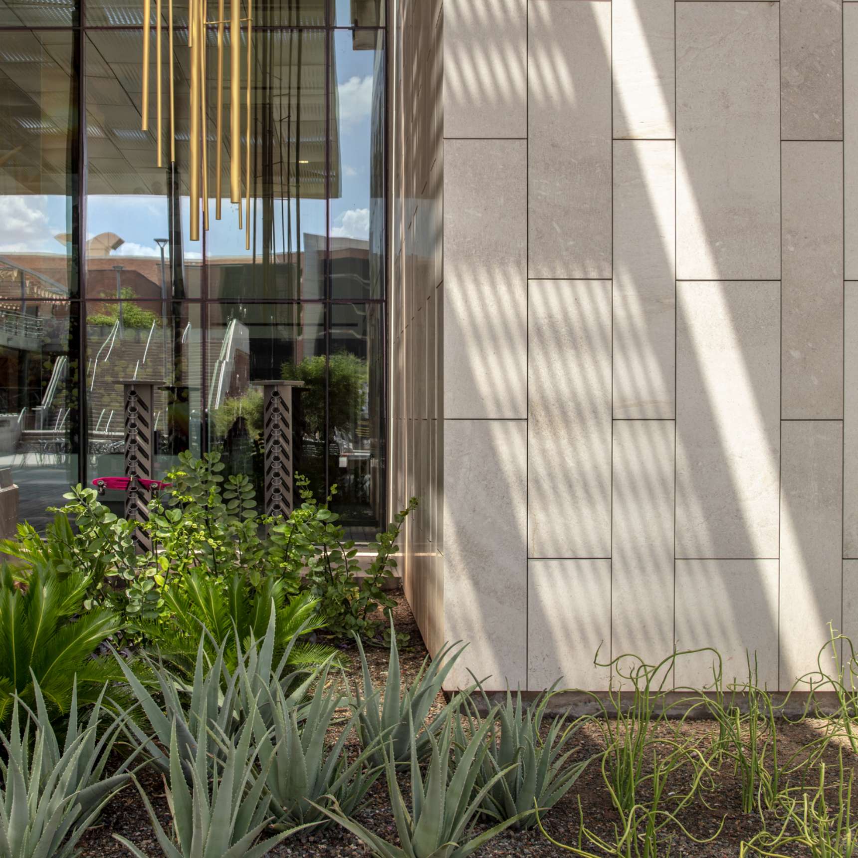 Modern building with beige stone facade, adjacent to a window reflecting vertical yellow light fixtures. The foreground has a landscaped garden with various green plants and succulents.