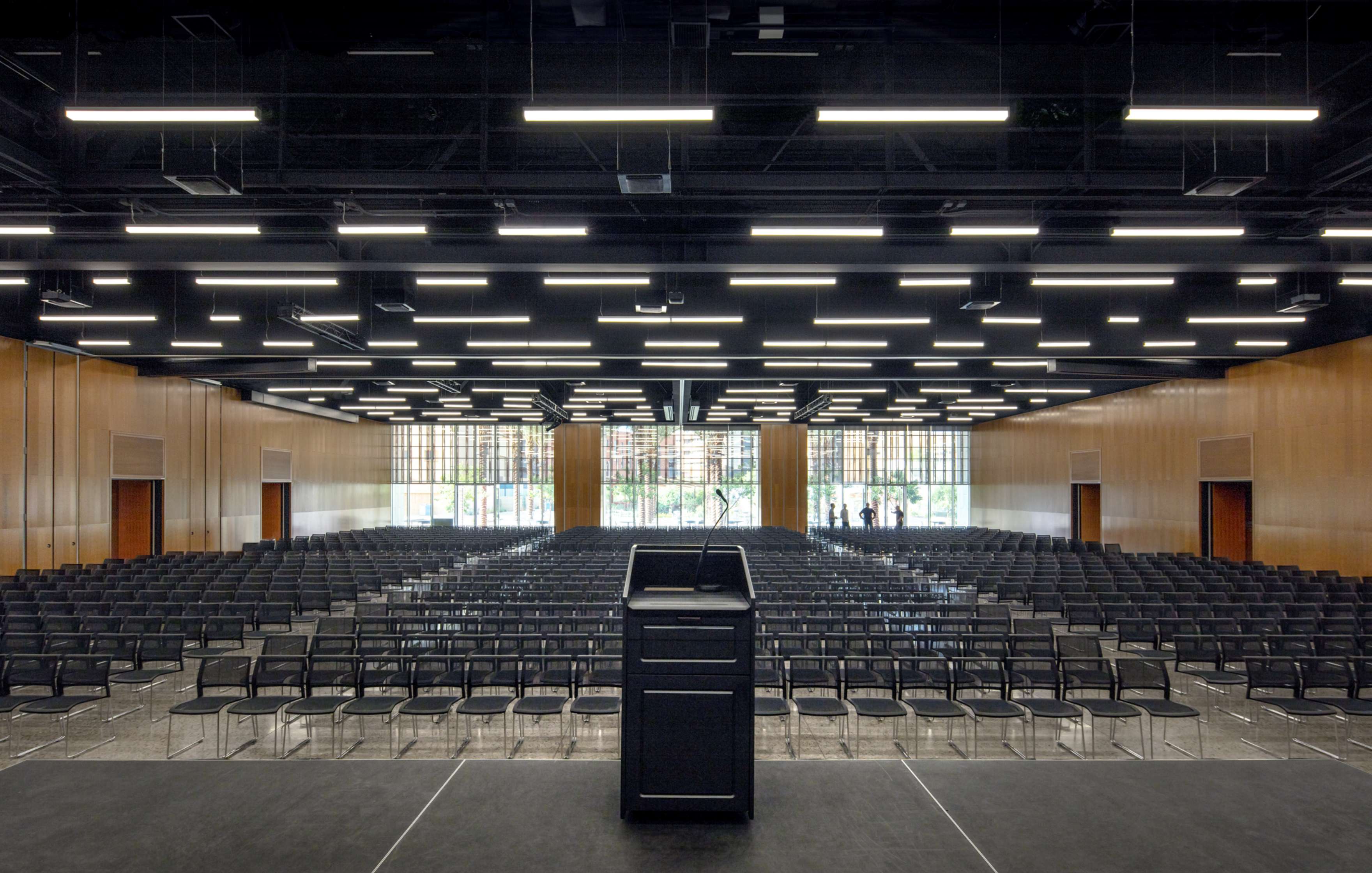 A modern conference hall with rows of empty chairs, a podium on stage, and large windows at the back letting in natural light.