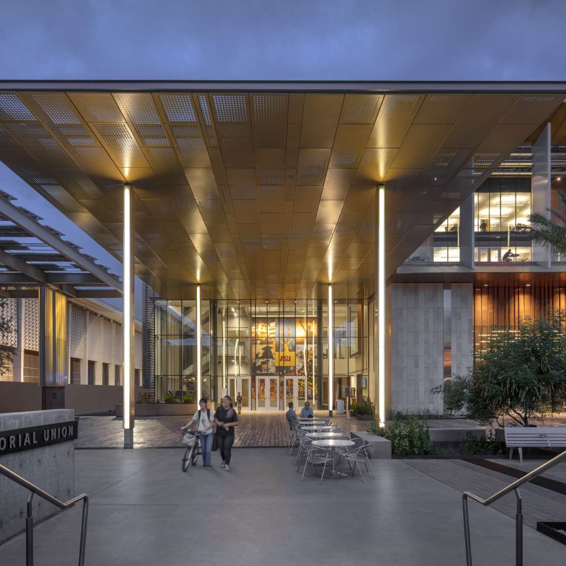 The image shows the modern exterior of a building with a large, illuminated canopy, glass entrance, and outdoor seating area with tables and chairs. Two people walk towards the entrance.