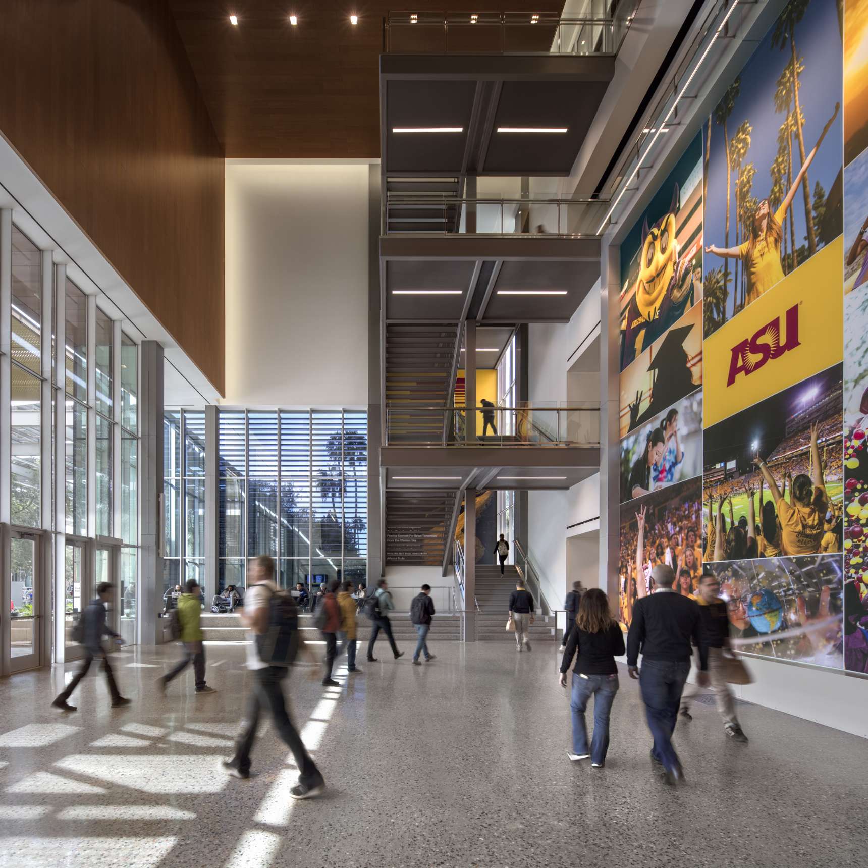 People walk through a spacious, modern building lobby with large windows, a staircase, and vibrant wall murals featuring the ASU logo and various images.