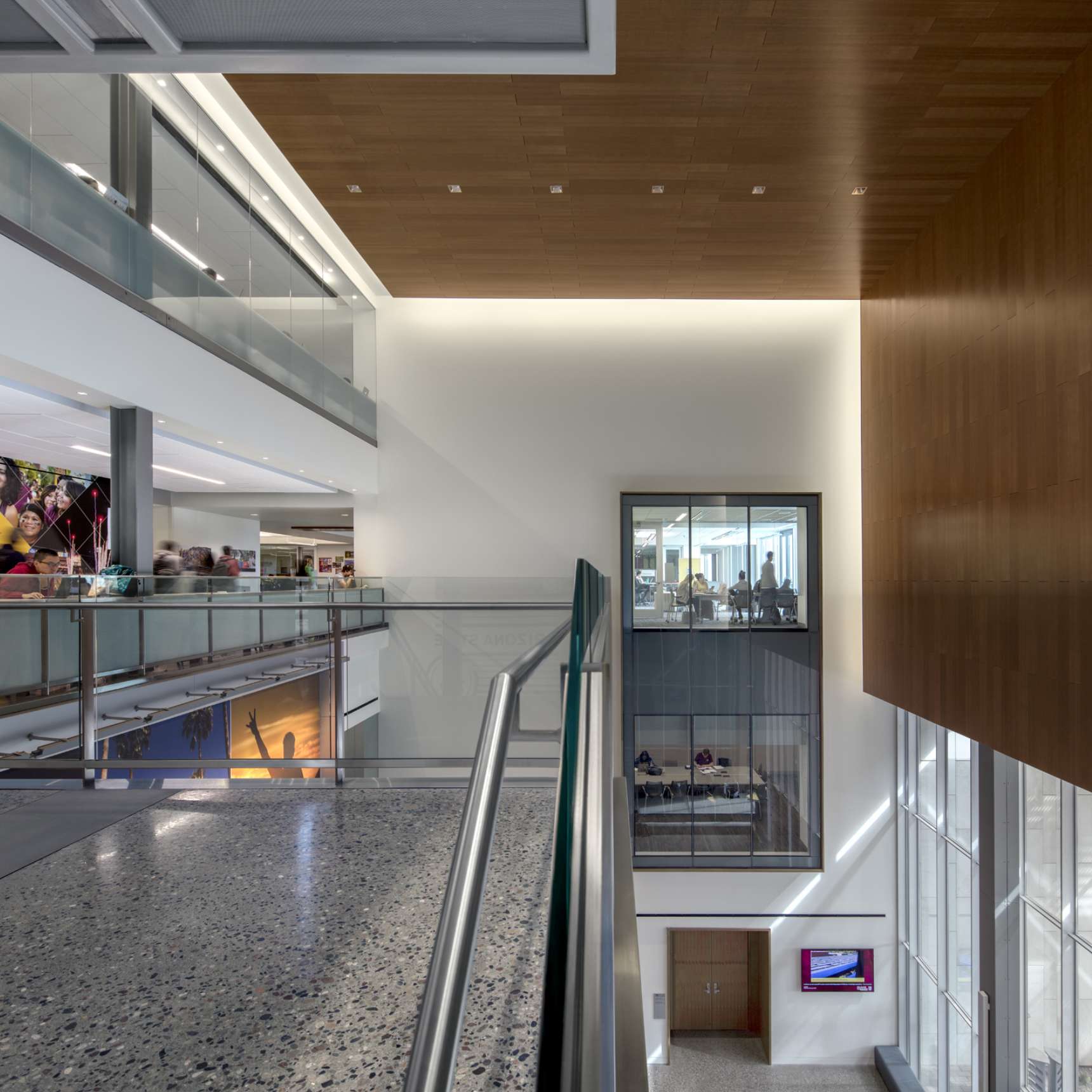 Modern office interior with multiple levels, glass railings, and a wooden accent wall. People are visible on the upper floor, and an office space is seen through a large window on the right.