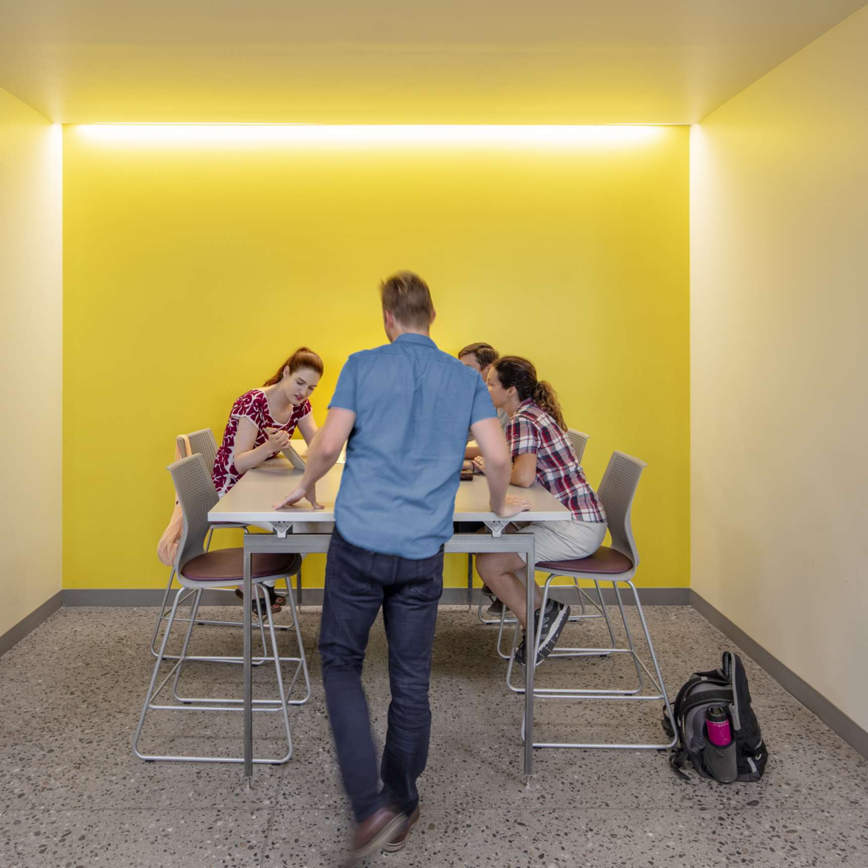 A group of people sit around a table with laptops and papers in a room with yellow walls, while one person stands and reaches towards the table. A backpack and water bottle are on the floor.