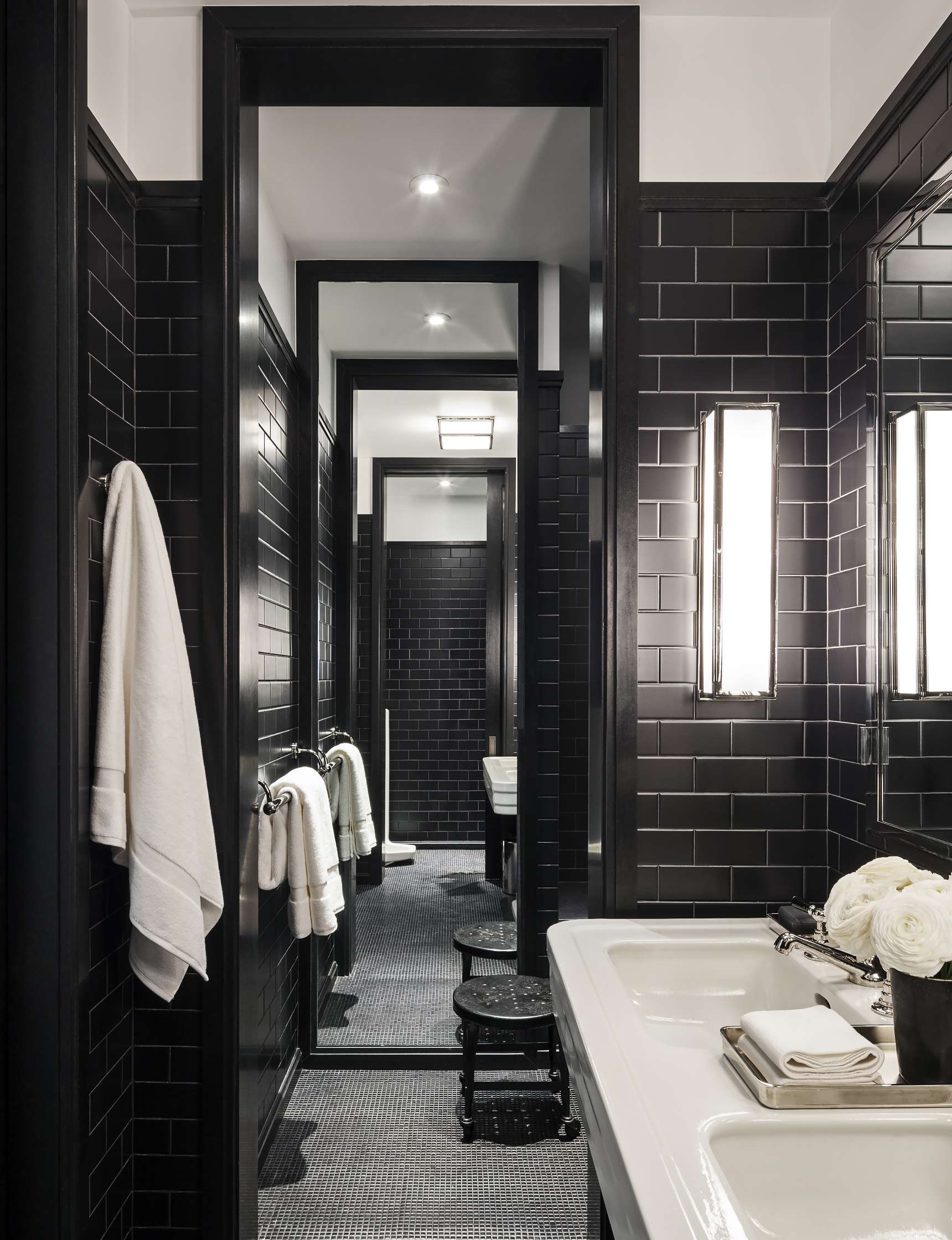 A modern bathroom features black subway tiles, a white sink with towels and flowers, and a hallway with multiple framed doorways leading to another room.