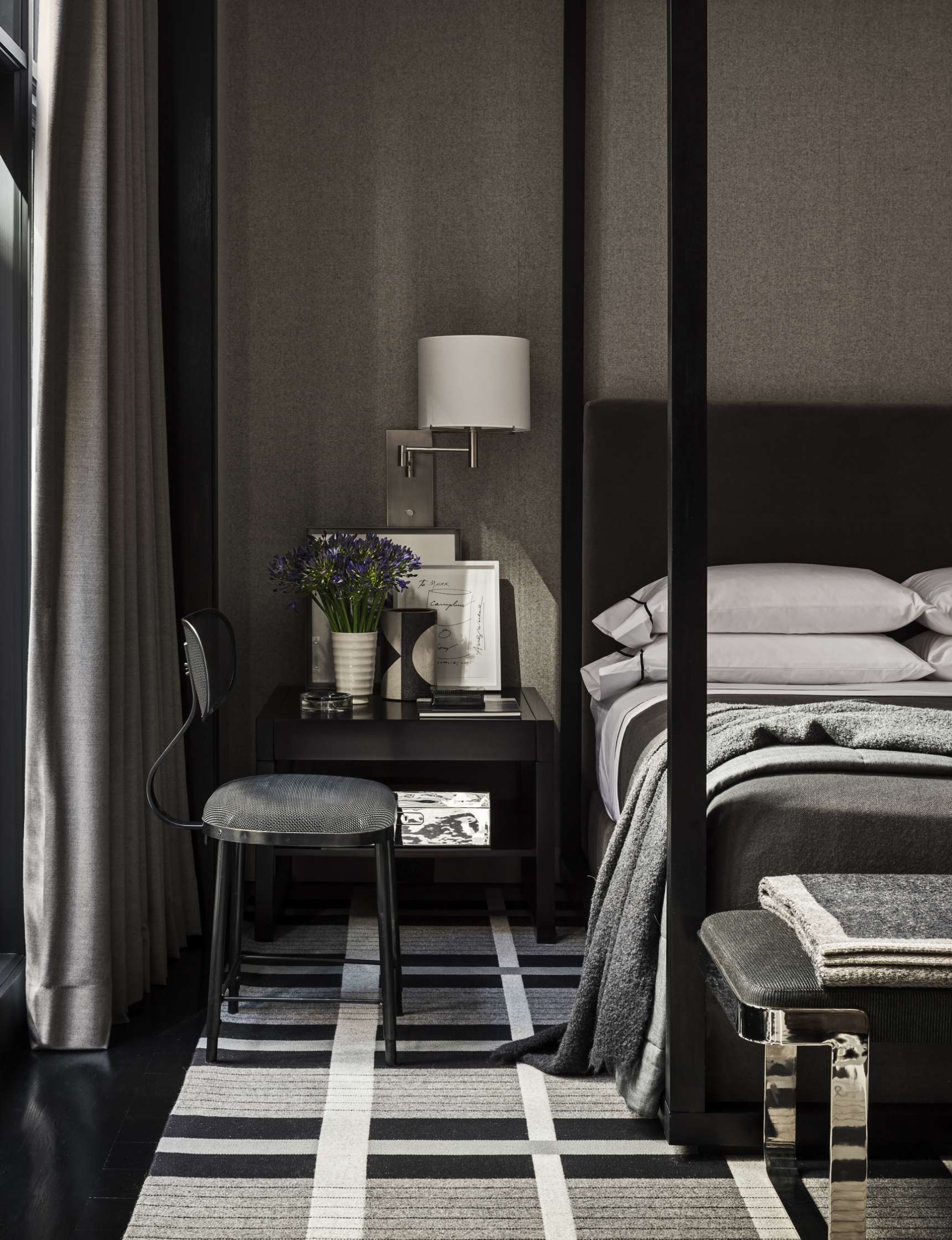 A modern bedroom with dark wood furniture, a side table with purple flowers, a wall lamp, books, a grey rug with stripes, and a neatly made bed featuring grey and white linens.