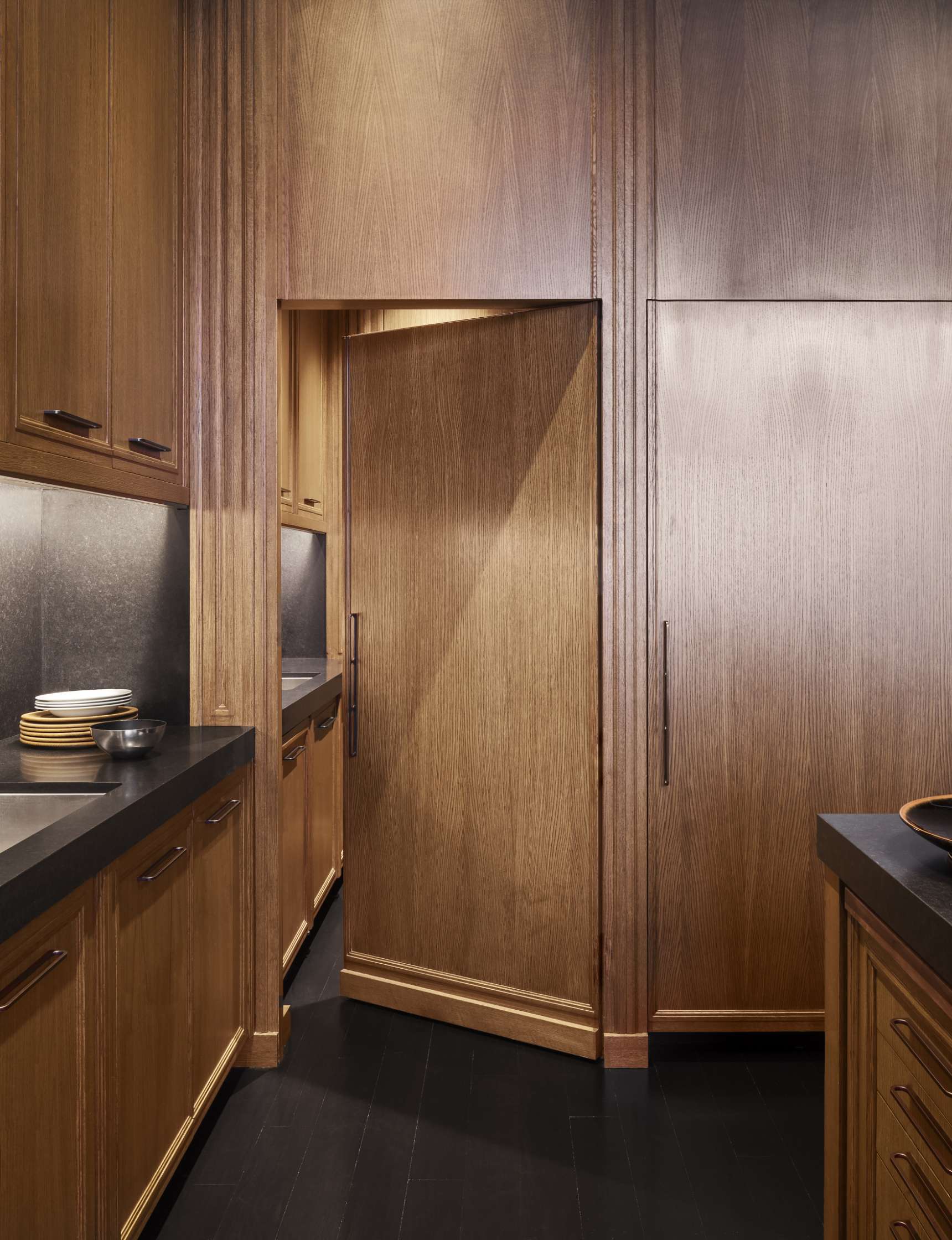 A modern kitchen with wooden cabinets, a central hidden door blending into the cabinetry, black countertops, and neatly arranged dishes.