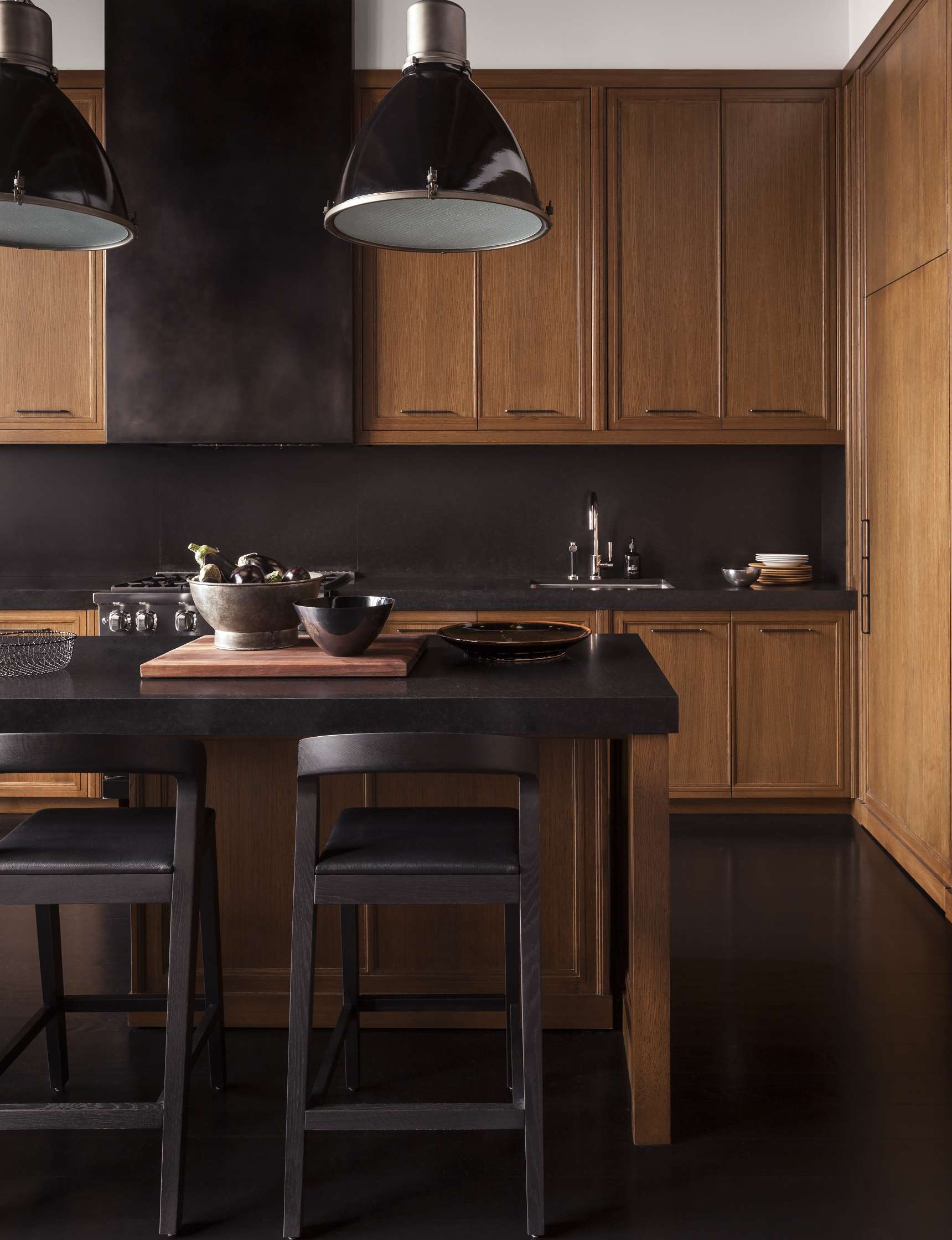 A modern kitchen with wooden cabinets, dark countertops, two black pendant lights, a central island with two black stools, and various kitchen utensils and bowls on the island.