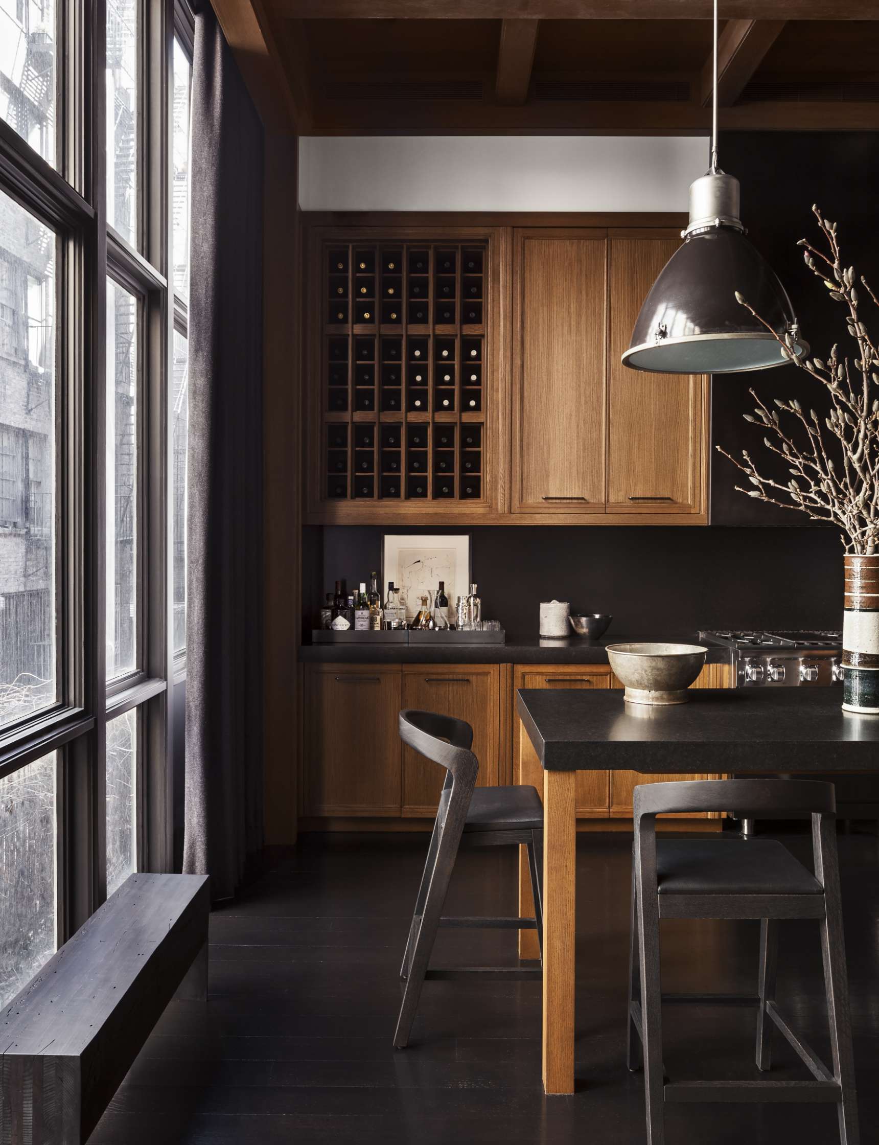 A modern kitchen with dark wood cabinets, a wine rack, a black countertop island with barstools, and a large window with long curtains. A black pendant light hangs above the island.