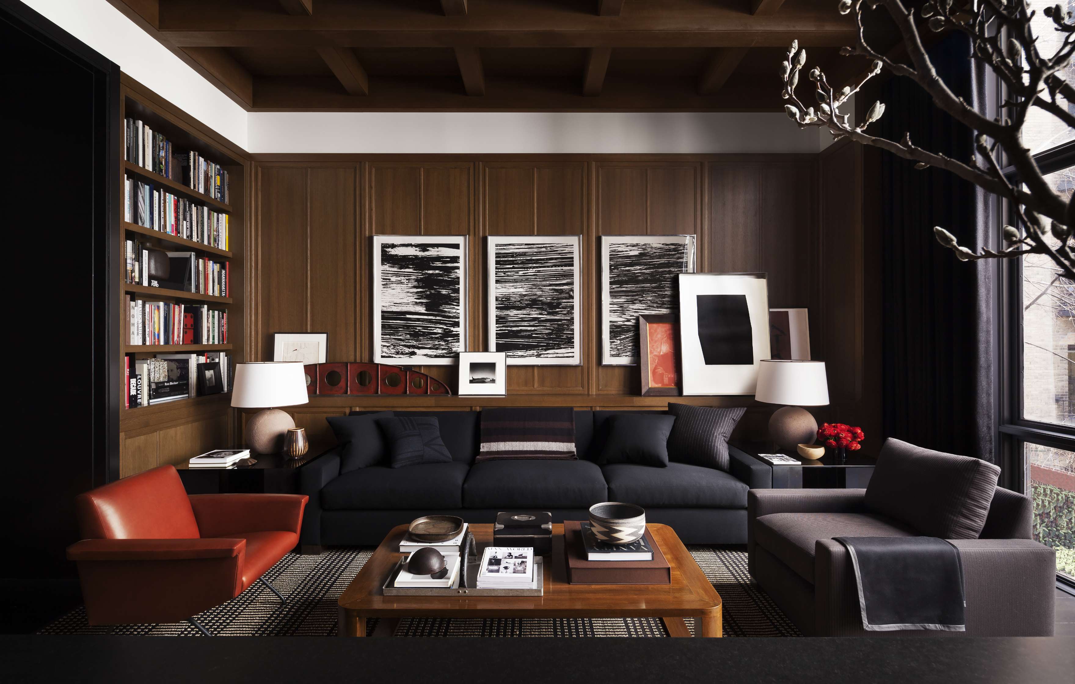 A modern living room with two dark sofas, an orange armchair, wooden coffee table, bookshelves, framed abstract art on the wall, and large windows with a view outside.