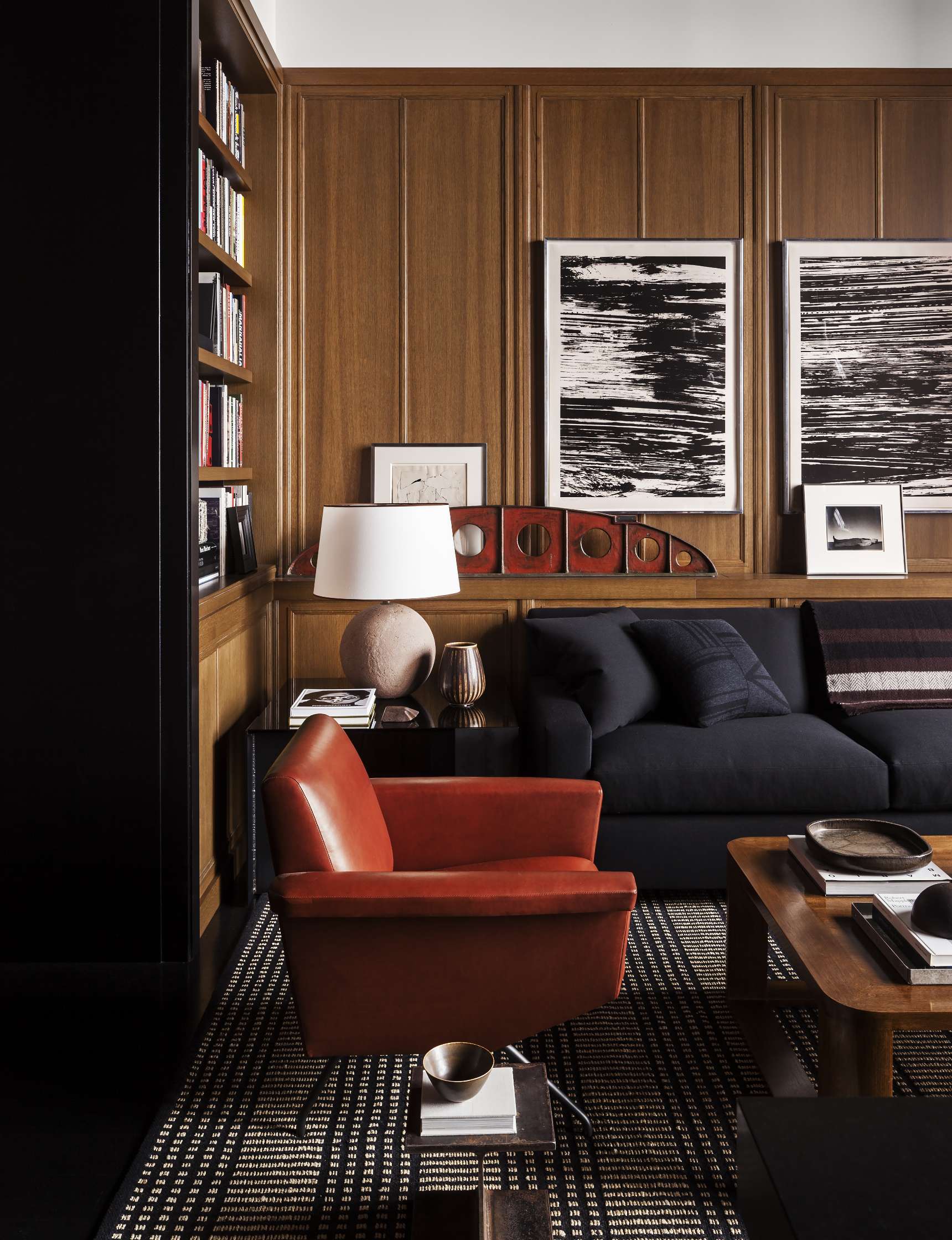 A modern living room with wooden panel walls, a black sofa, a red armchair, black-and-white abstract art, and bookshelves. A table lamp and various decor items are present.