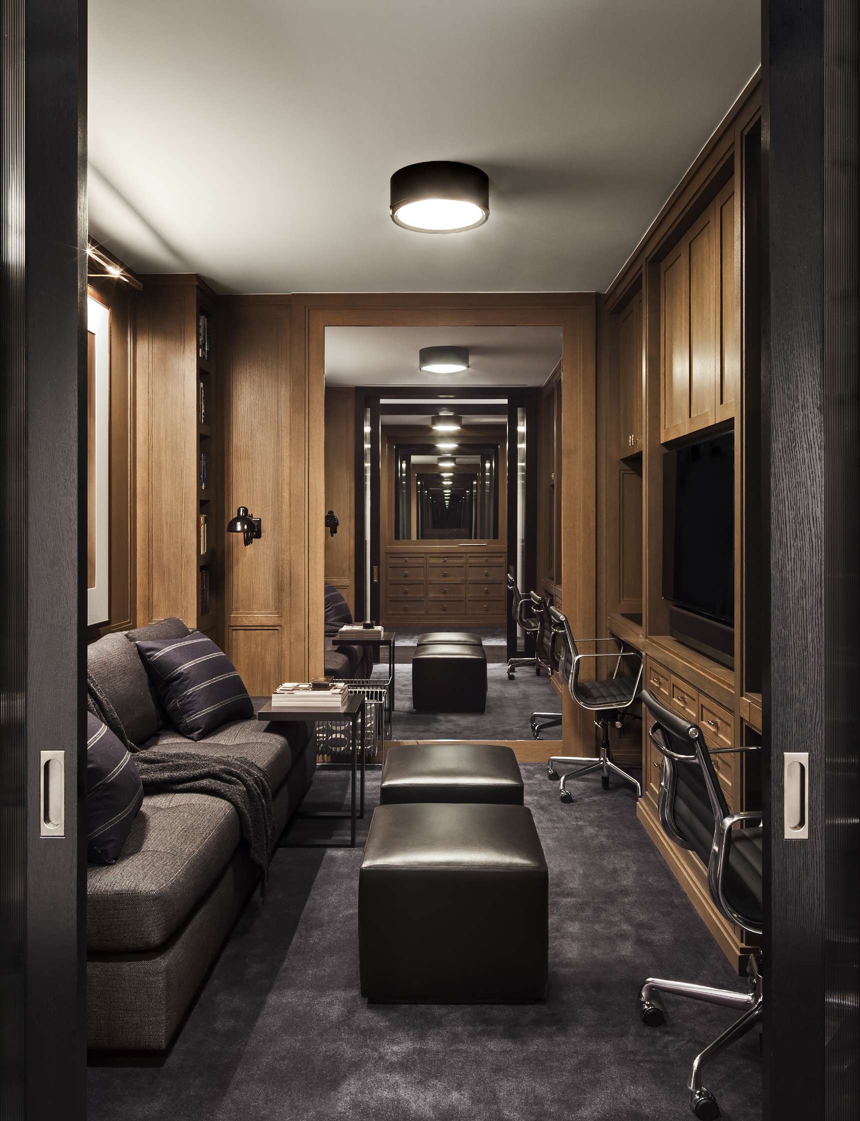 A modern office room featuring a sofa, two ottomans, a coffee table, several chairs, a flat-screen TV, and wood-paneled walls. There are also mirrored walls creating an illusion of extended space.