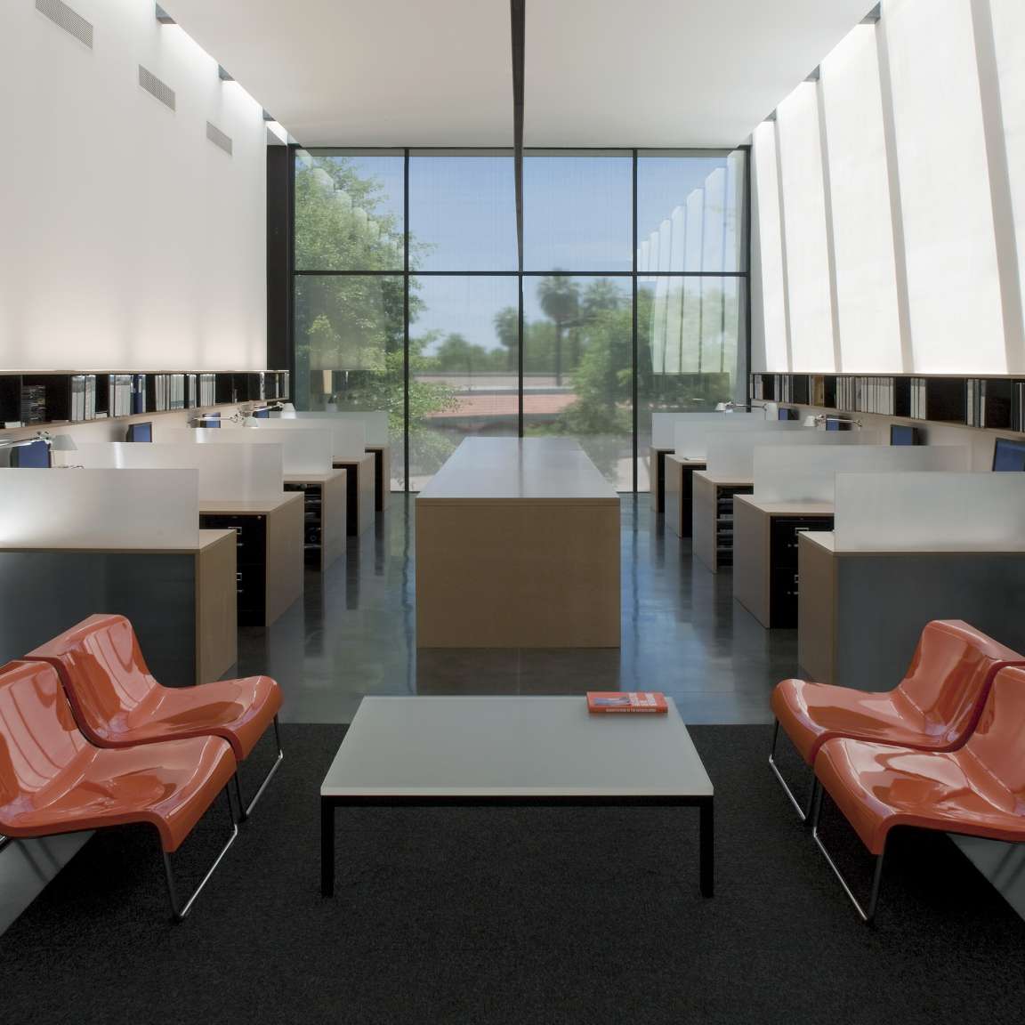 A modern office space with two rows of workstations along both walls, two orange chairs, a coffee table in the foreground, and large windows at the far end letting in natural light.