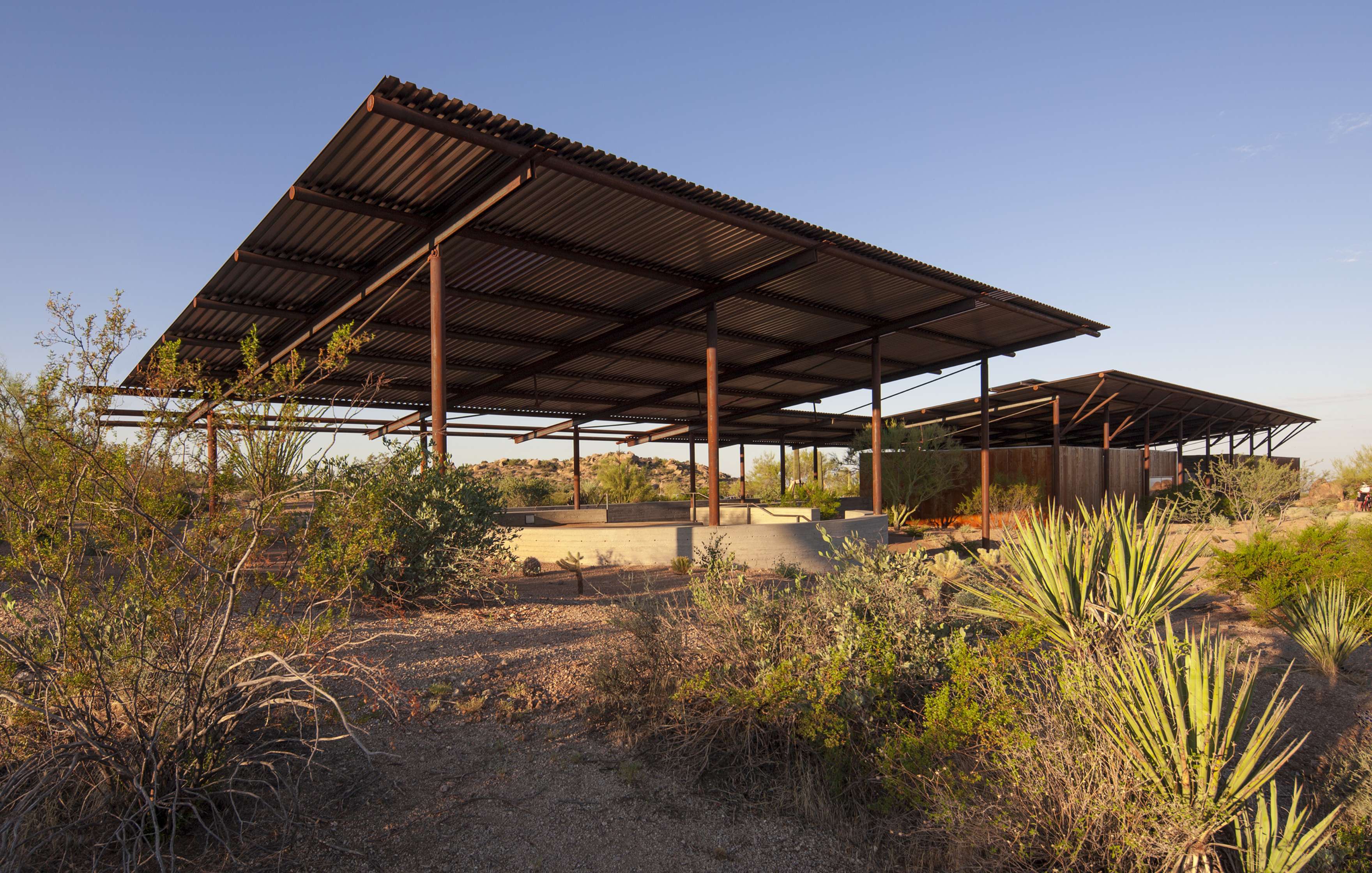 Covered outdoor structures with an open framework, situated in a desert landscape with various desert plants in the foreground.