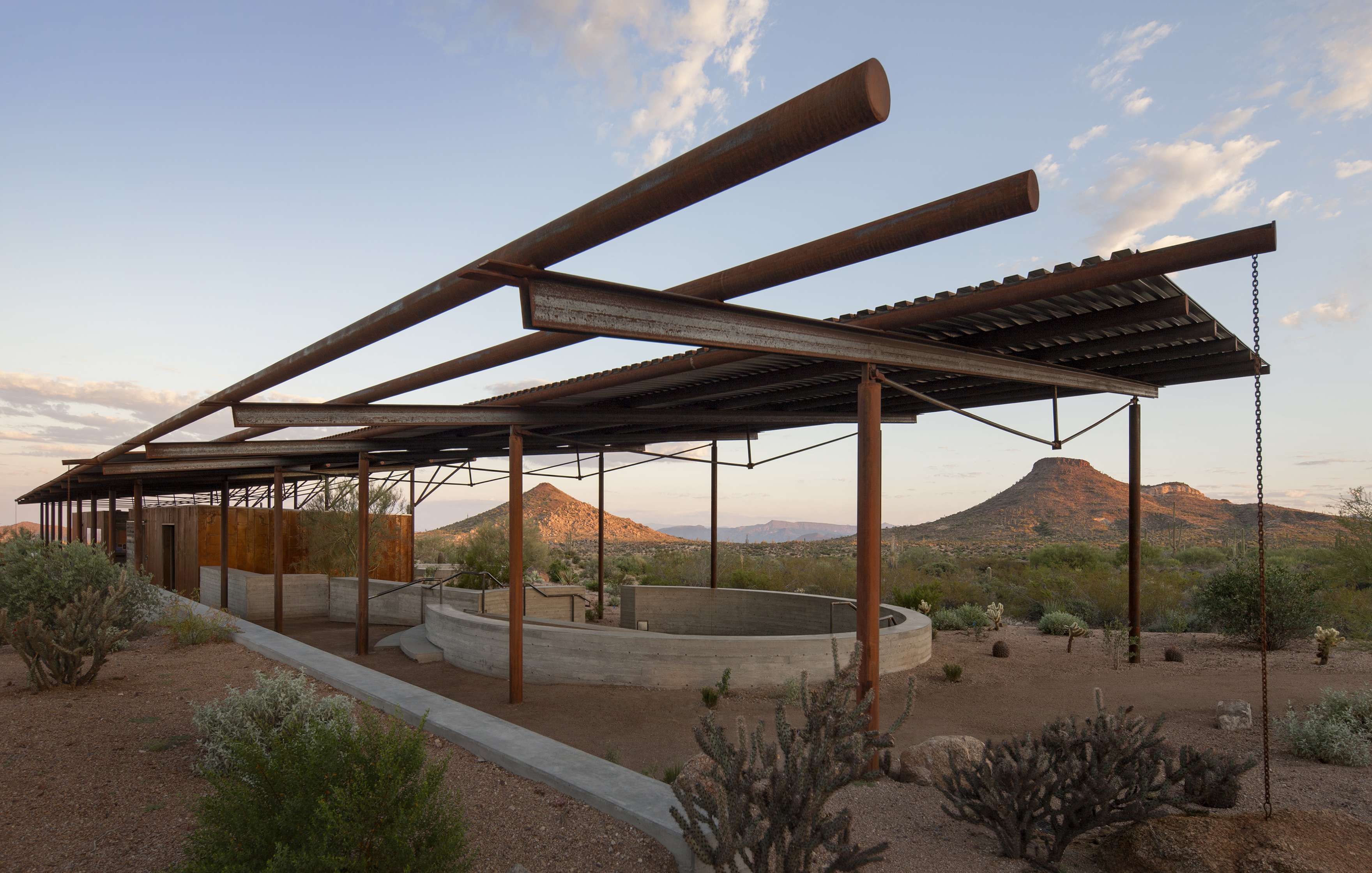 A structure with an open roof made of metal beams and corrugated sheets stands in a desert landscape with shrubs and distant mountains in the background.