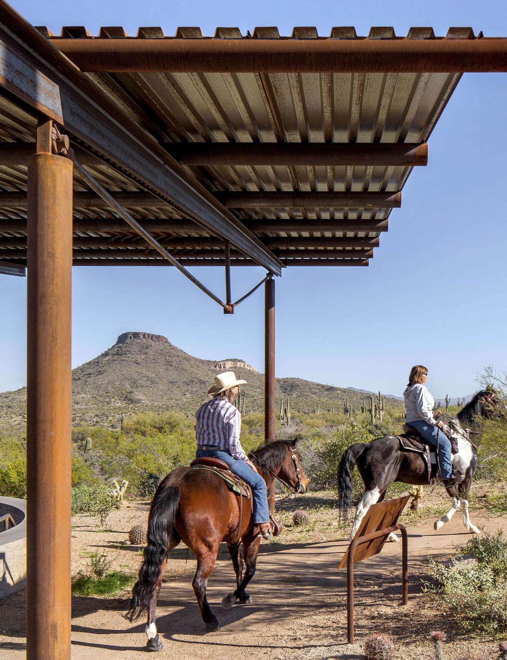 Two people riding horses in a desert landscape with a mountain in the background, under a corrugated metal shelter. Cacti and desert vegetation surround the area.