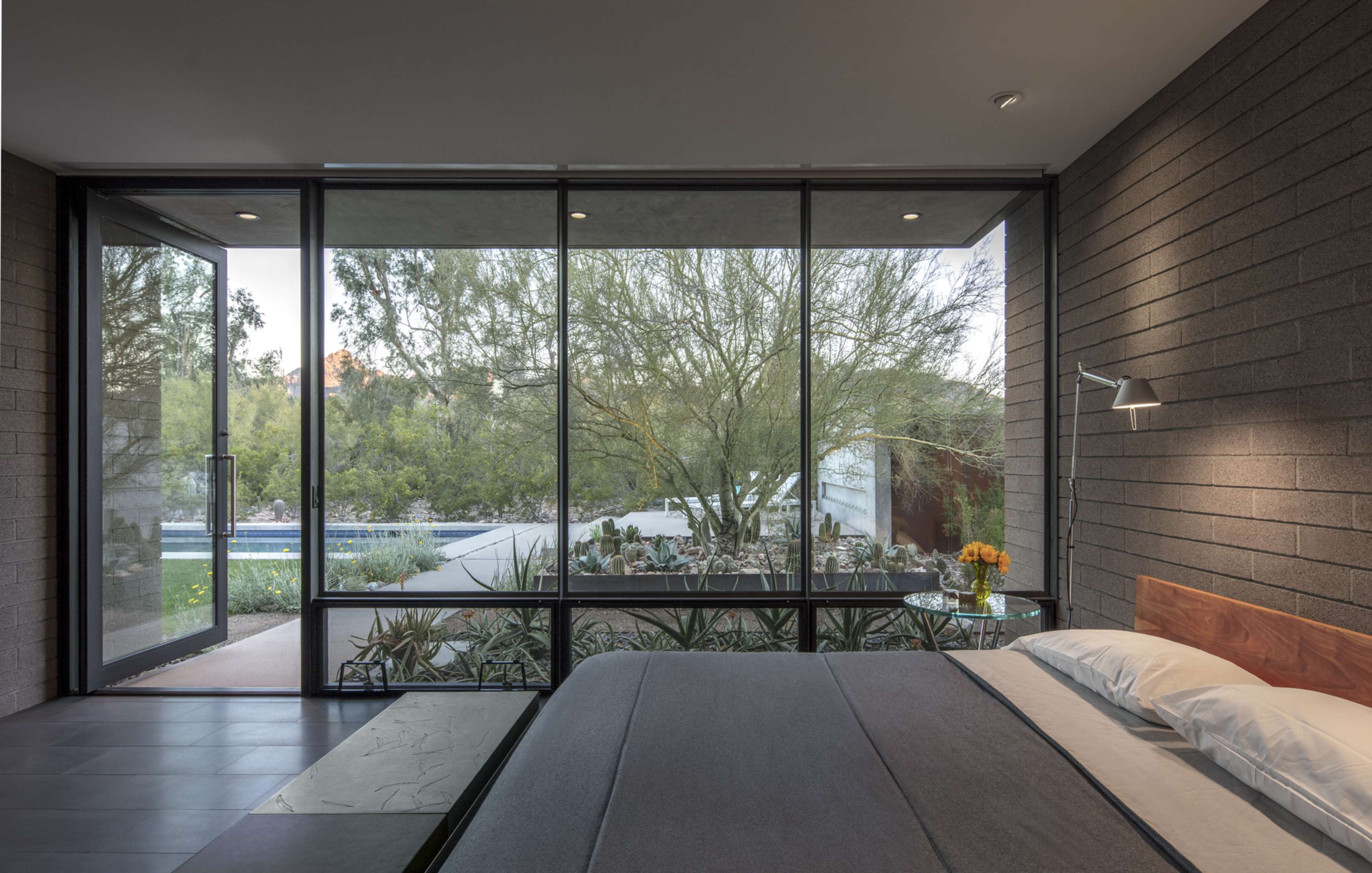 Modern bedroom with large floor-to-ceiling windows overlooking a garden with trees and a swimming pool. The room has a neatly made bed, a wall lamp, and a stone-textured wall.