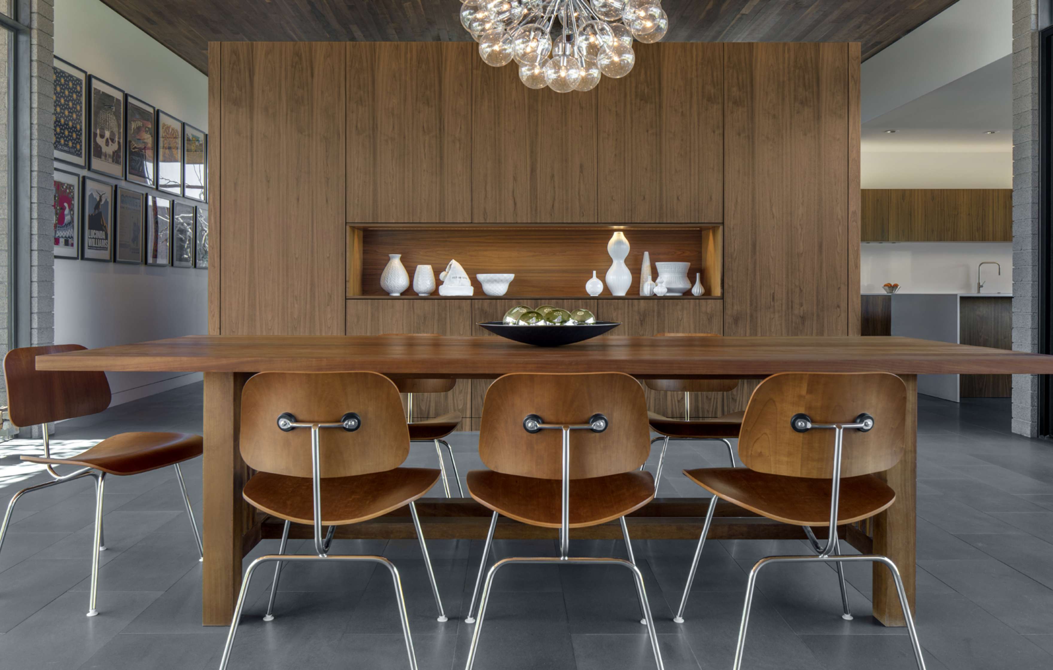 A modern dining room with a wooden table and six brown chairs. A bowl of green apples is on the table, and the wooden wall decorates with shelves displaying white vases. A chandelier hangs above.