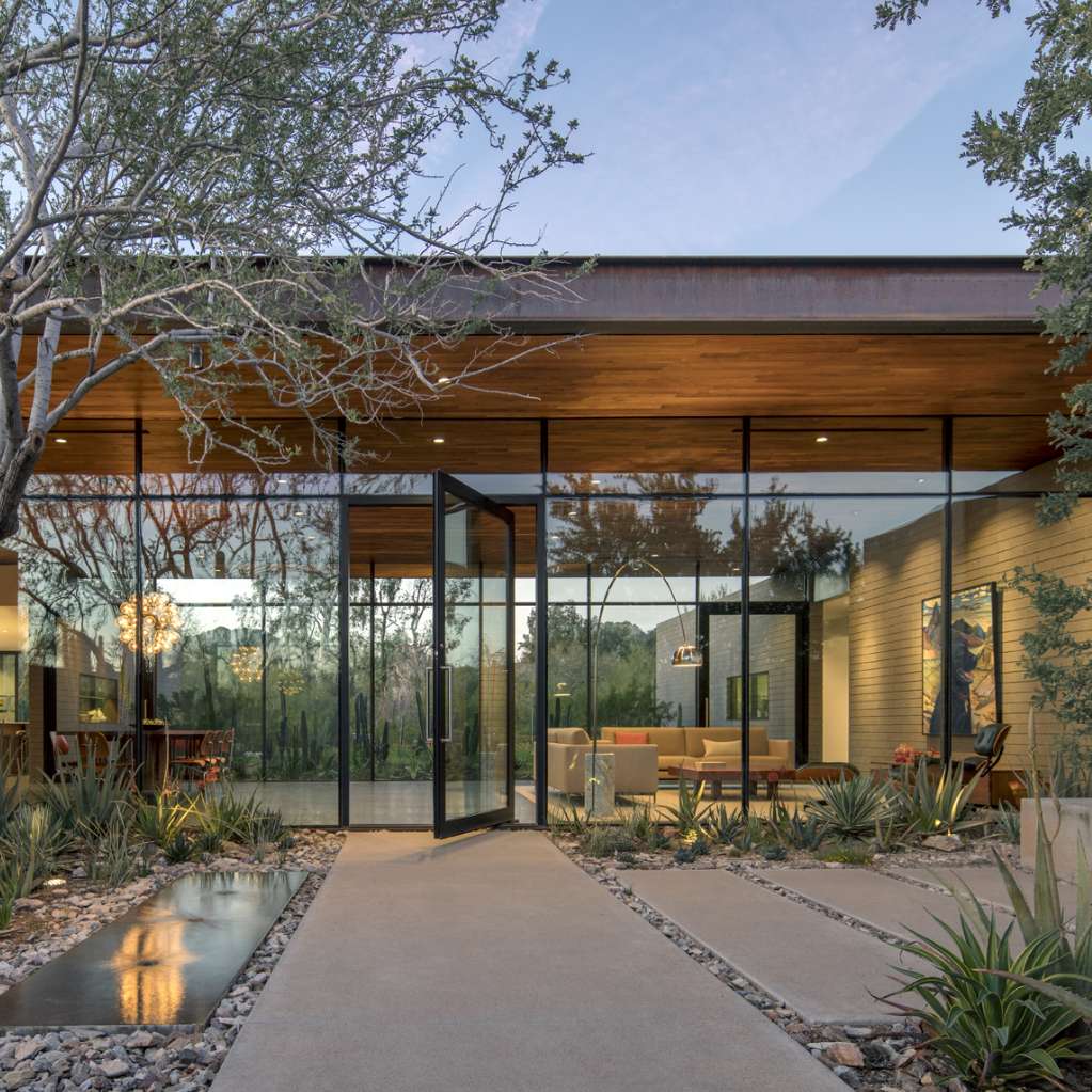 Modern house exterior with large glass windows, a wooden roof, and minimalist landscaping featuring trees, shrubs, and a concrete path. The interior is visible through the glass, showing modern furnishings.