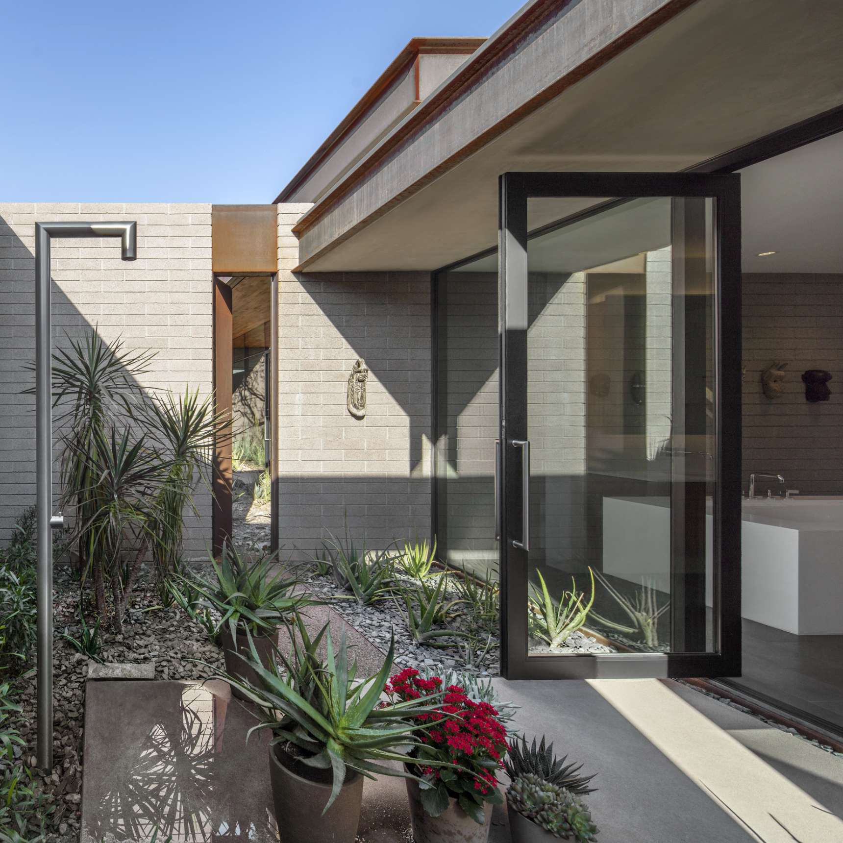 Modern indoor-outdoor bathroom area with large glass door, various potted plants, succulents, stone path, and a mix of natural and artificial light.