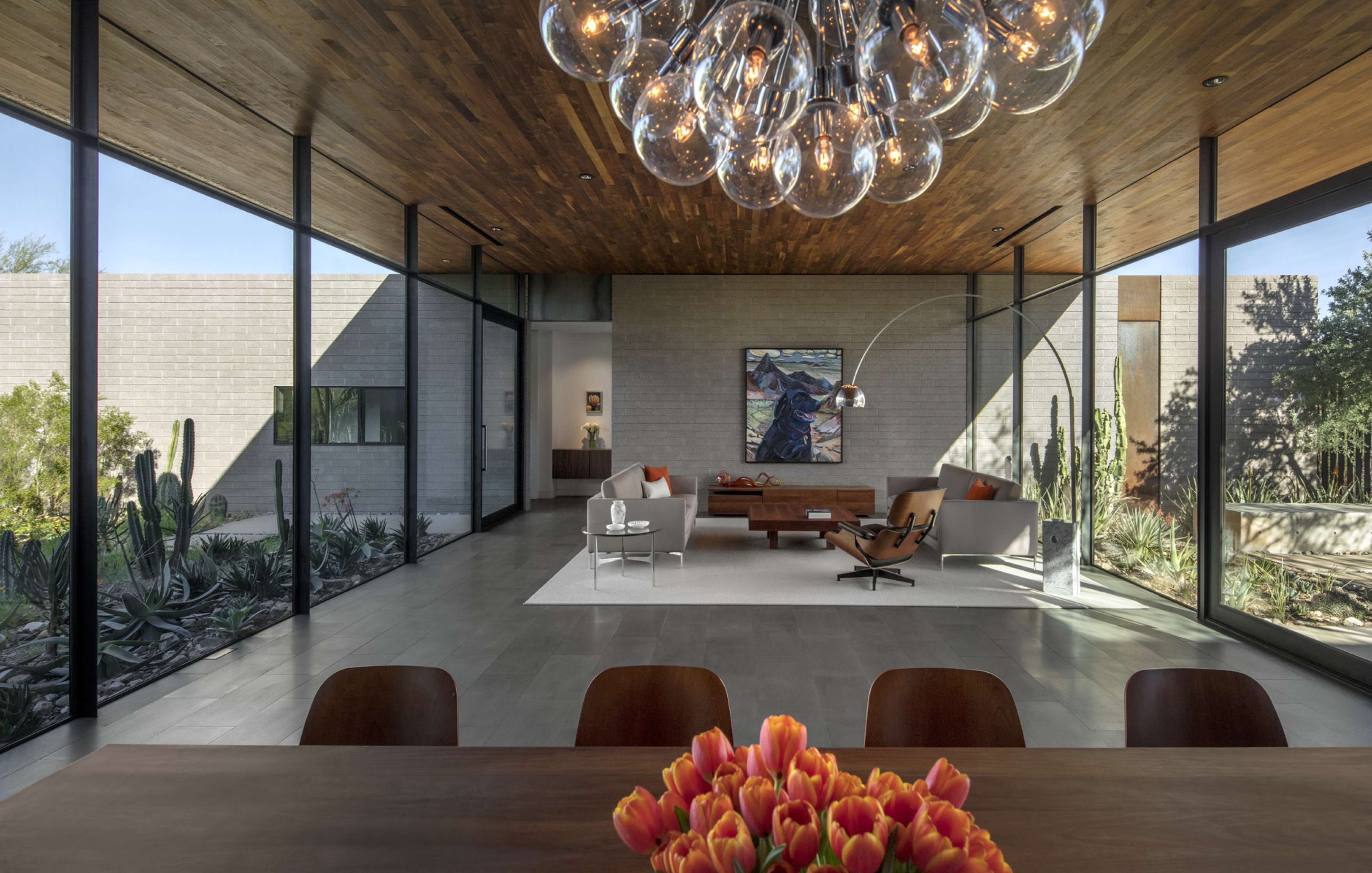 Modern living room with large windows, a wooden ceiling, and a chandelier. Furnishings include a sofa, armchairs, and a dining table with tulips. The room opens to an outdoor garden area.