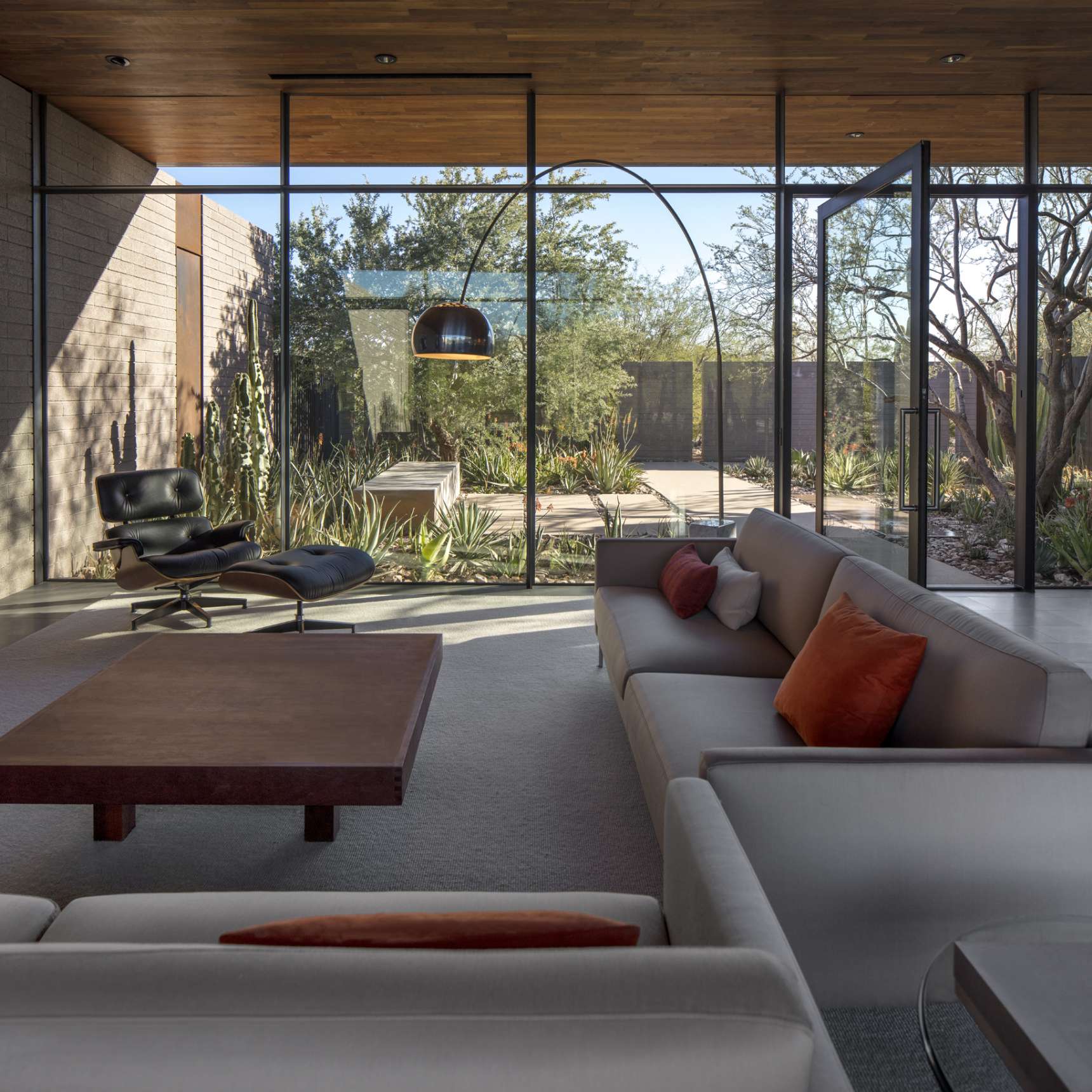 Modern living room with large floor-to-ceiling windows, a low square coffee table, white sectional sofa with orange pillows, and an Eames lounge chair with a view of a landscaped patio.