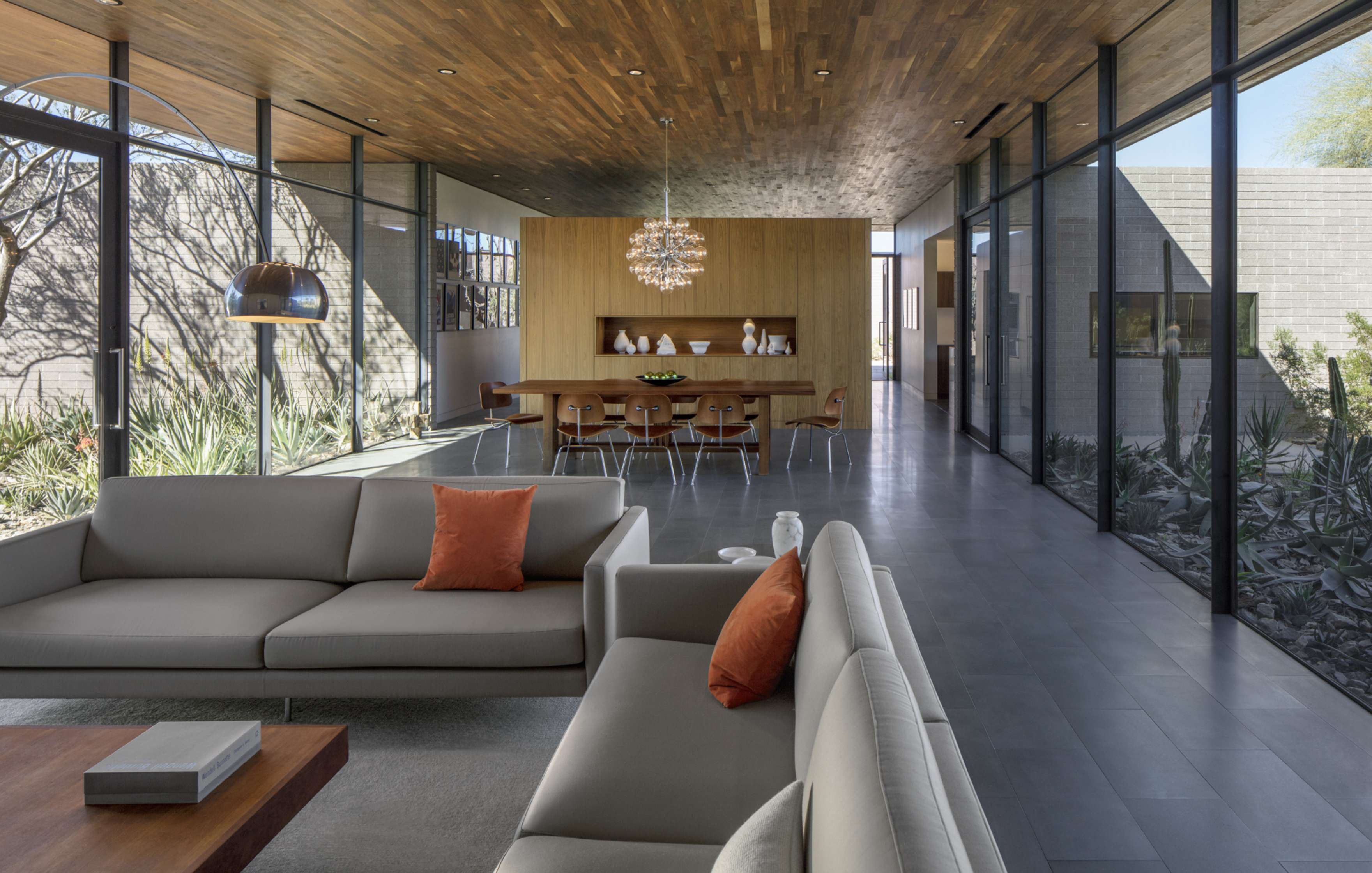 Modern open-concept living and dining area with floor-to-ceiling windows, grey sofas with orange pillows, a chandelier, wooden ceiling, and garden views.