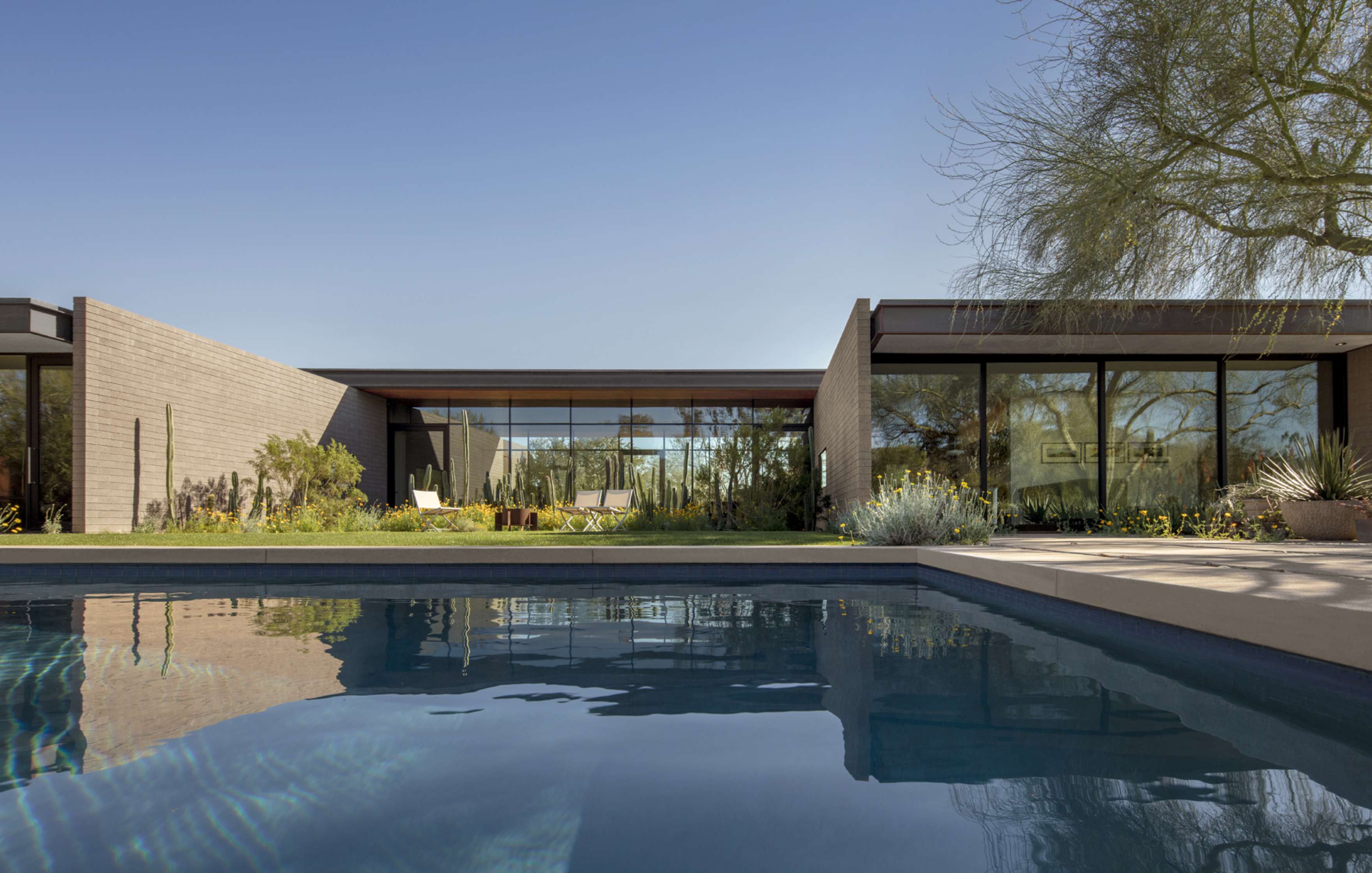 Modern single-story house with large glass windows, situated near a swimming pool, surrounded by desert landscaping and clear blue sky.