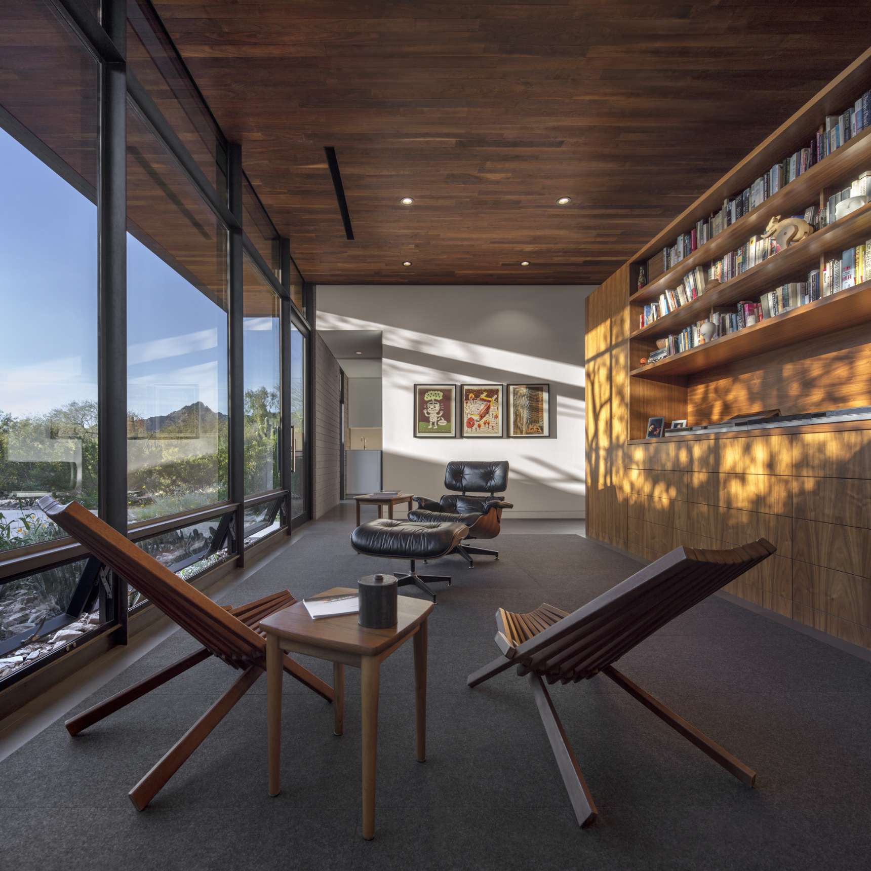 Contemporary sitting area with wood-paneled ceiling, large windows, lounge chairs, small table, and built-in bookshelves filled with books. Sunlight streams through the windows.