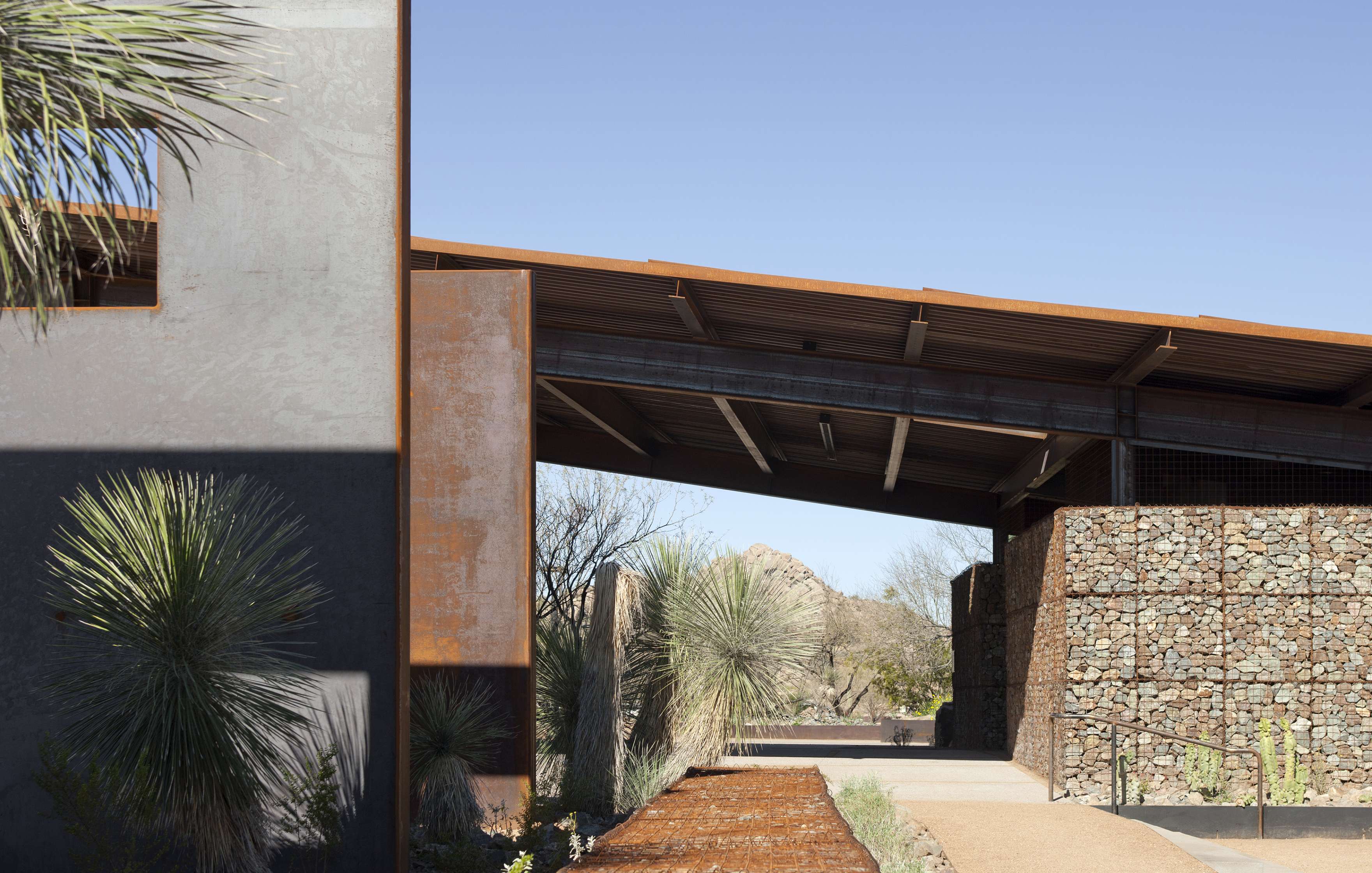 Modern building exterior with a mix of concrete, metal, and stone elements. The structure features an open roofed walkway under clear skies, with desert plants on each side.