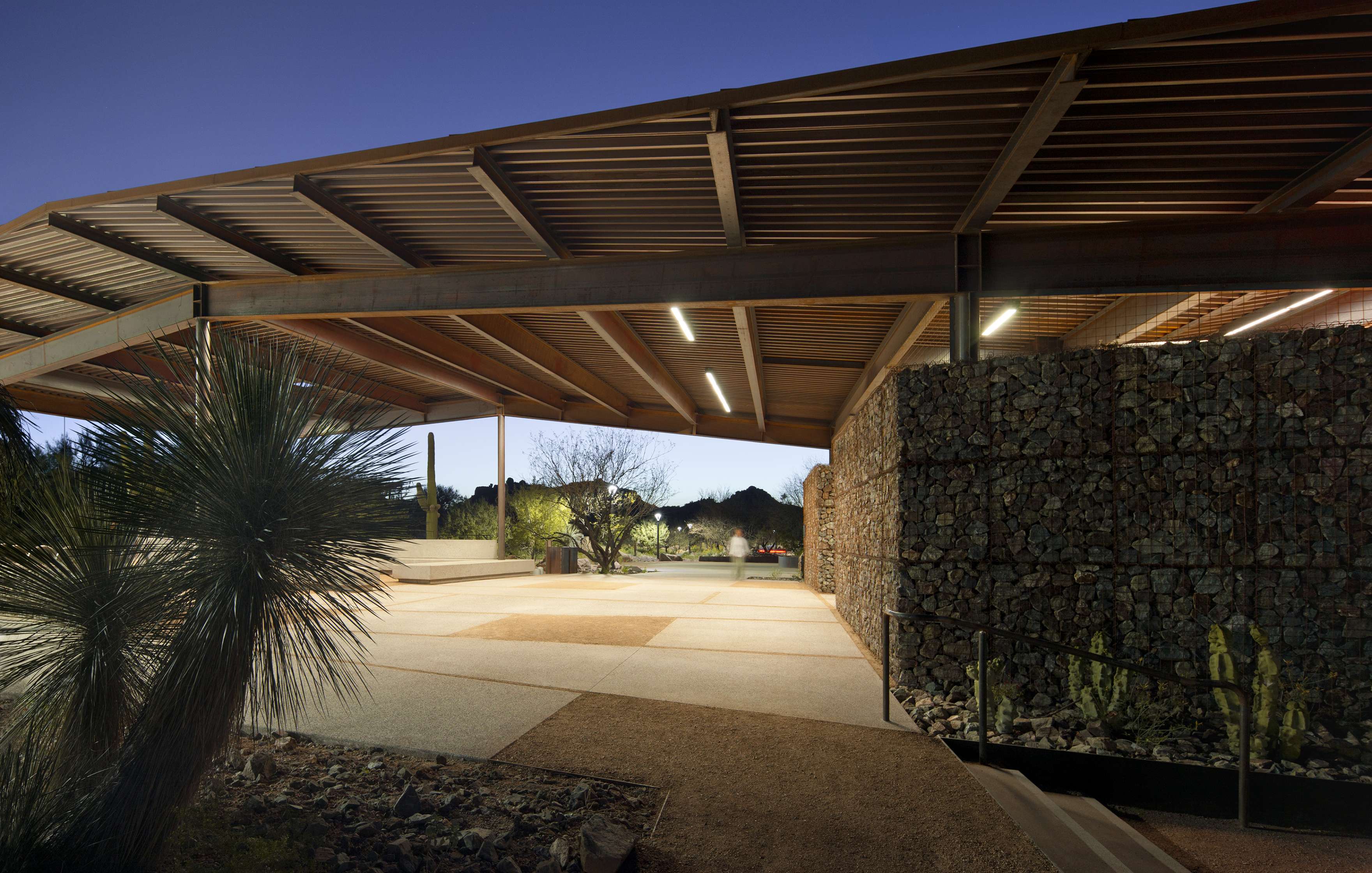 A modern outdoor pavilion with a metal roof and gabion walls at dusk, surrounded by desert landscaping and equipped with built-in seating areas.