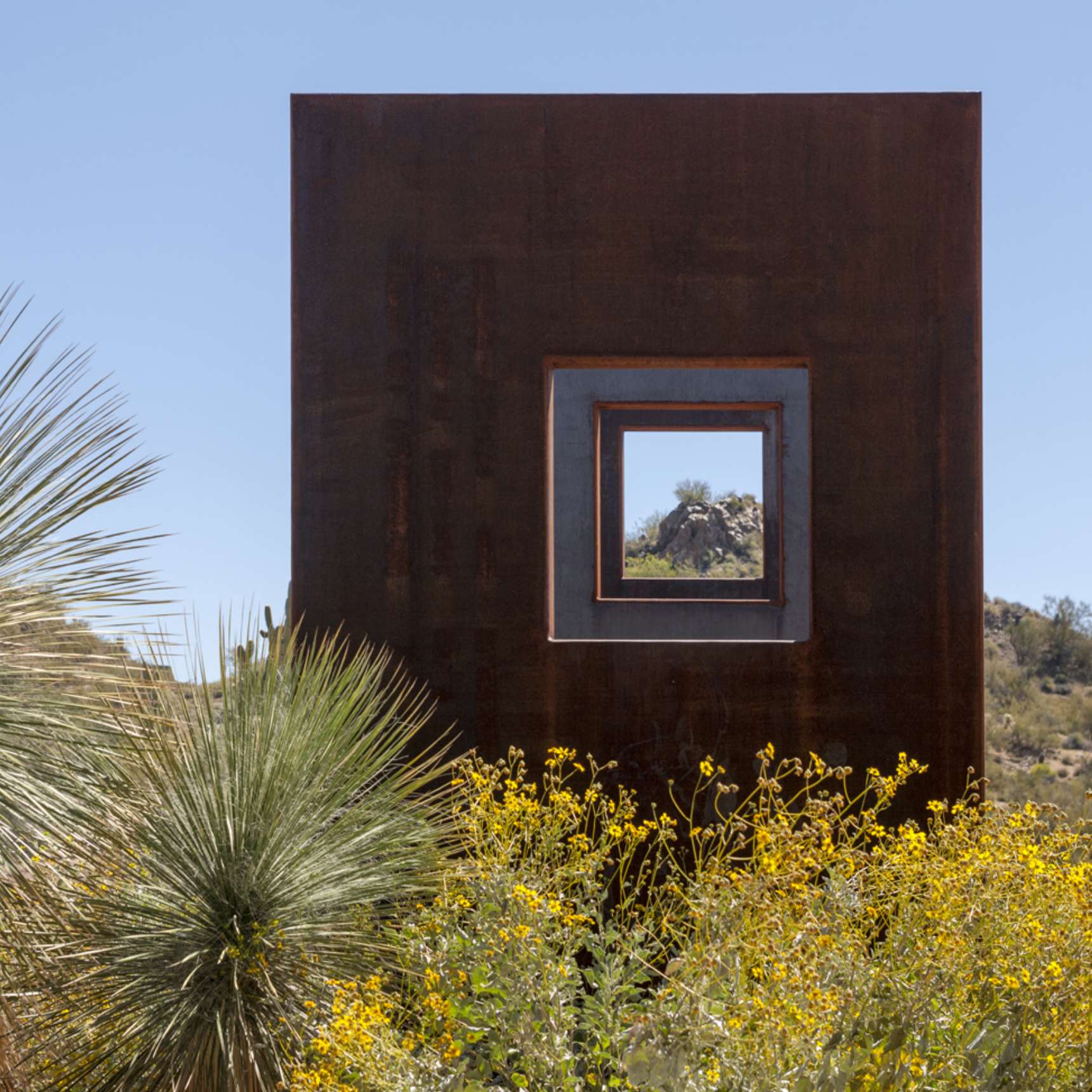 A rust-colored metal structure with square cutouts frames a desert landscape, including plants and a clear sky visible through the openings.