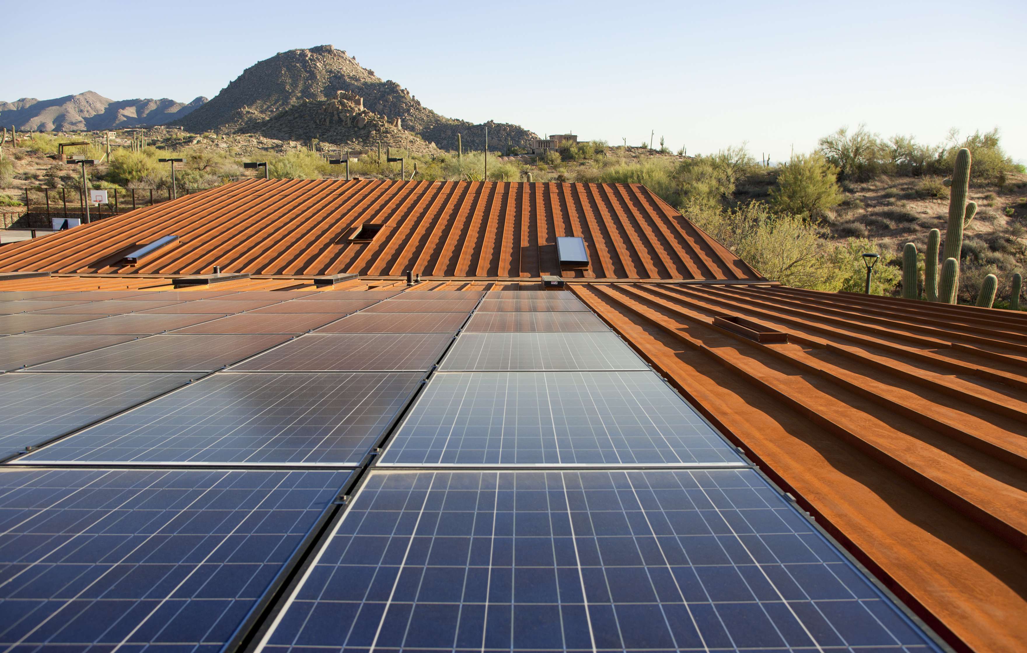 Solar panels installed on a rust-colored roof with desert landscape and a mountain in the background, under clear skies.