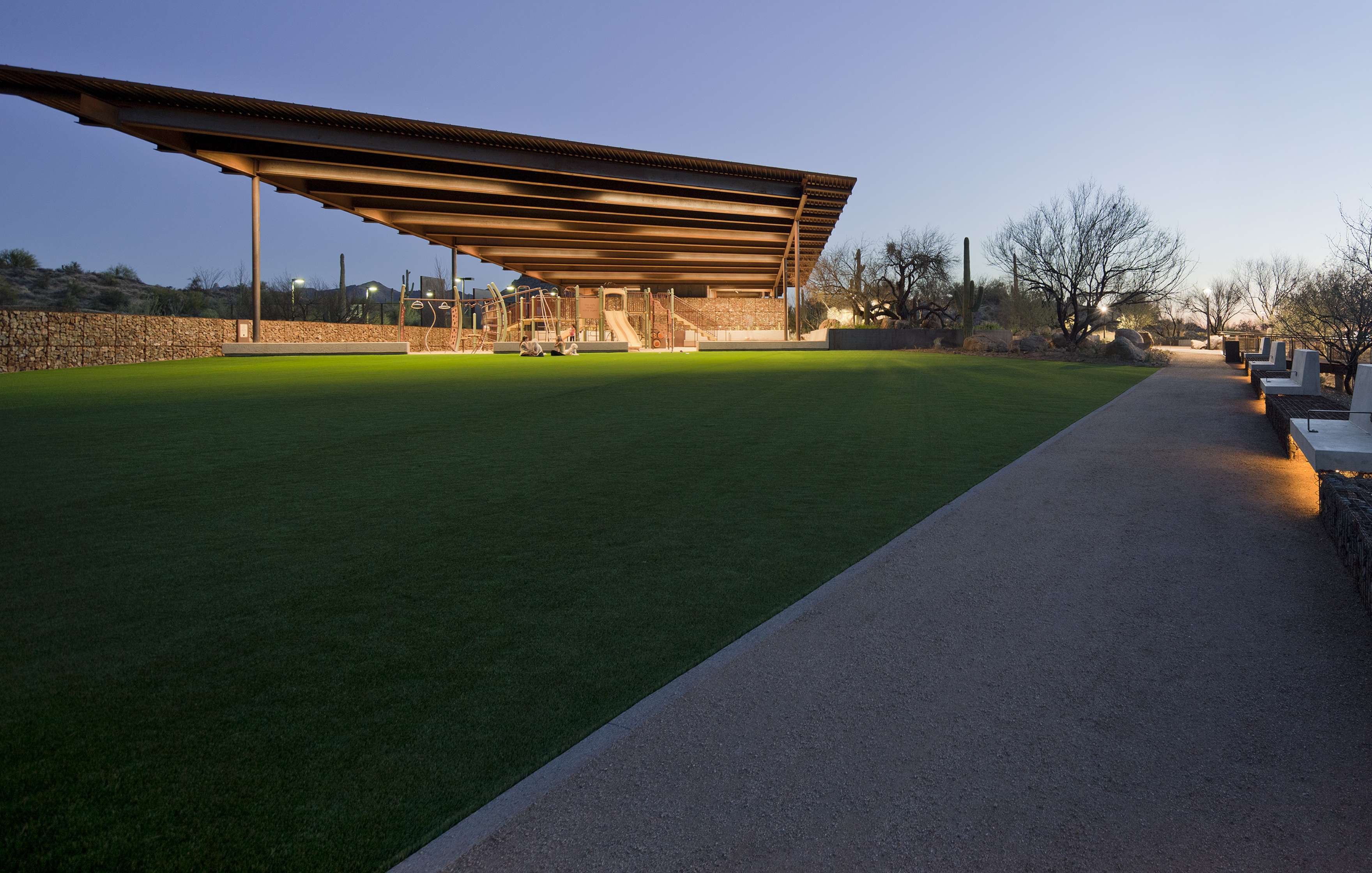 A modern architectural structure with a large open canopy covers a playground on a vast green lawn, adjacent to a paved pathway in a desert landscape at dusk.