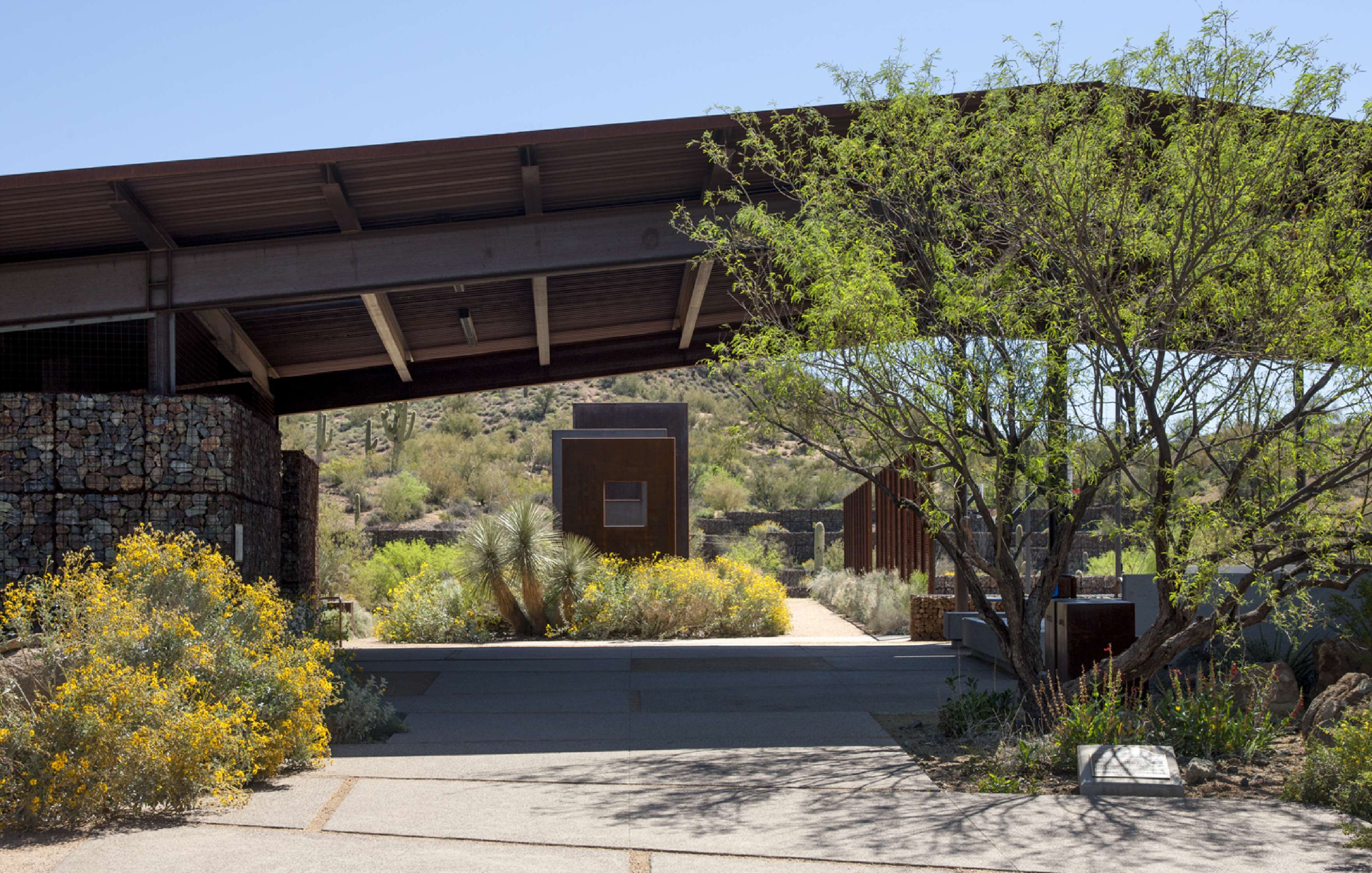 A modern structure with a large overhanging roof sits amidst desert vegetation, featuring flowering bushes and a few small trees, with a pathway leading to a wooden door in the background.