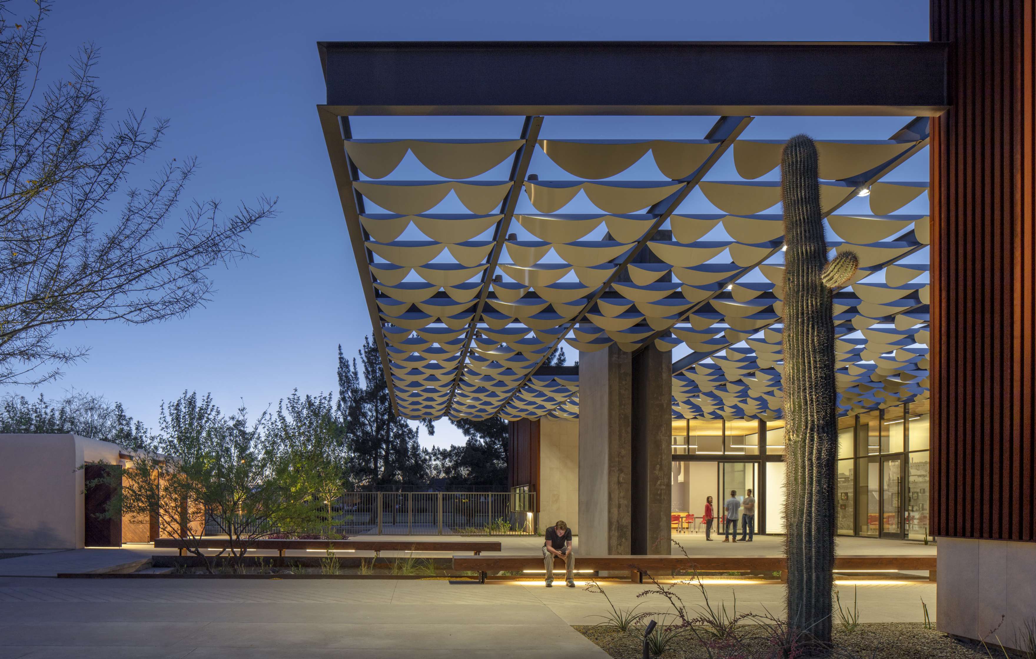 A modern building with geometric canopy, large windows, and seating area. A person sits on a bench near a cactus in the foreground, under a twilight sky.