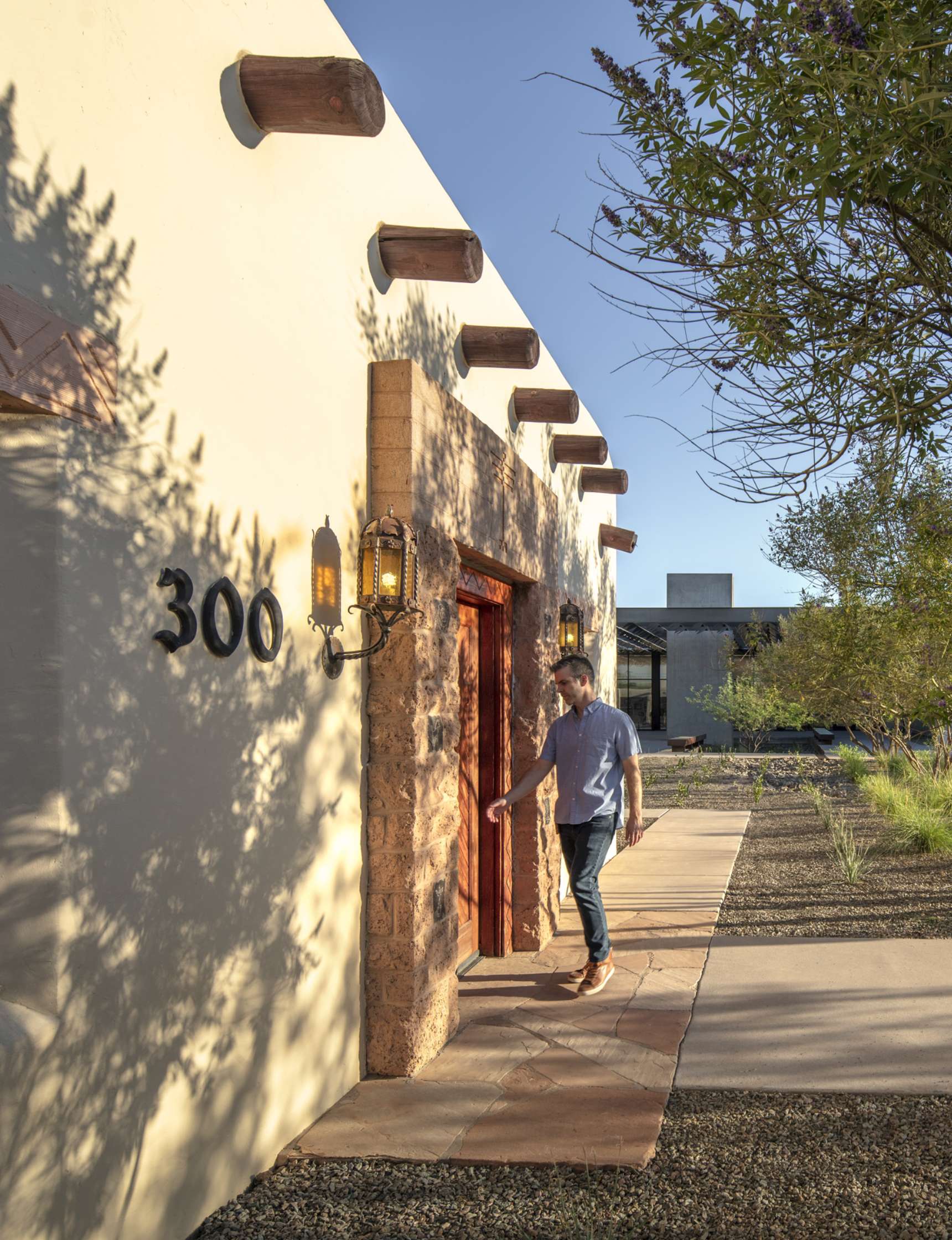 A person exits a building with the number 300 on the wall, under a lantern-style light fixture. The surrounding area includes trees and a path in an outdoor setting.