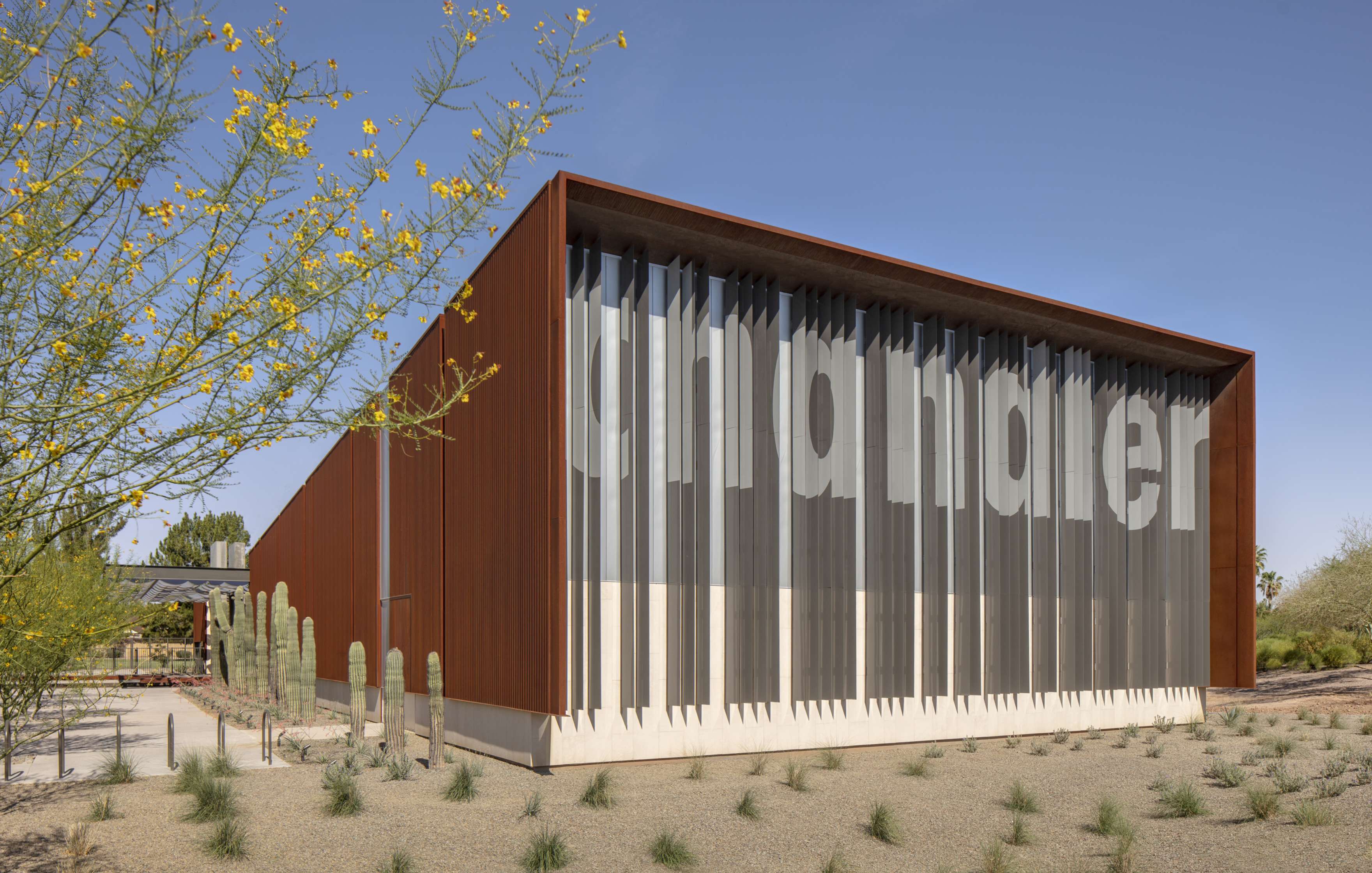 A modern building with a rust-colored exterior and vertical lines displaying the word “Chandler” stands in a desert landscape with cacti and sparse vegetation under a clear sky.