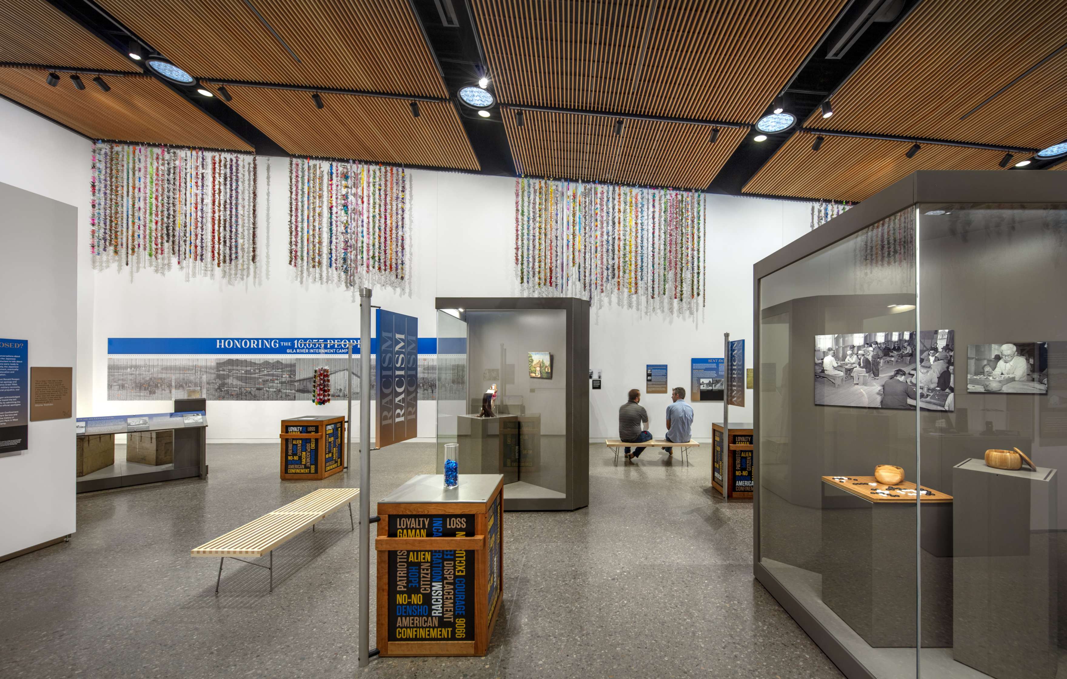 A modern exhibition hall with various displays, informational panels, and two seated people. The ceiling is adorned with hanging art pieces made from multicolored strips.