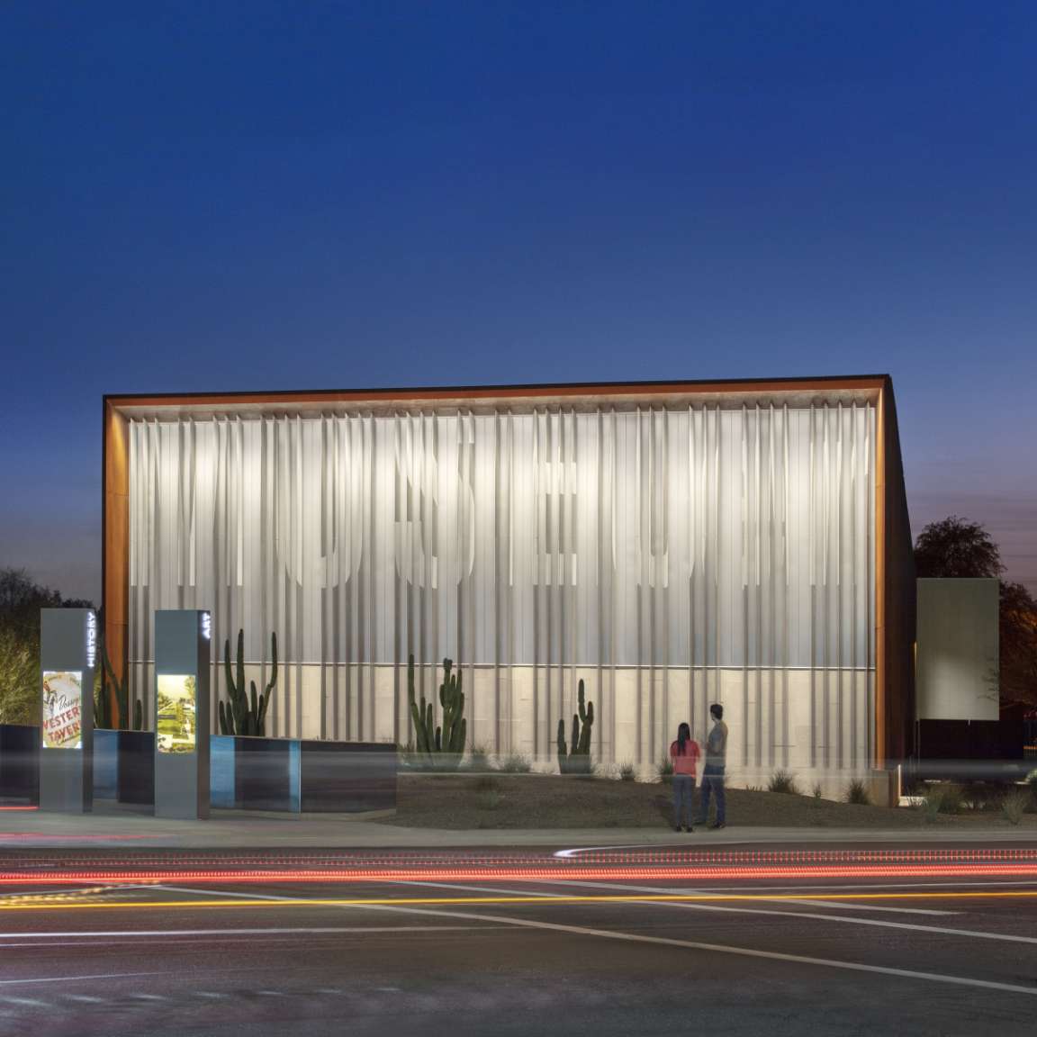 A museum with a modern facade and prominently lit signage is shown at dusk. Two people stand near the entrance, and light trails from passing vehicles are visible in the foreground.