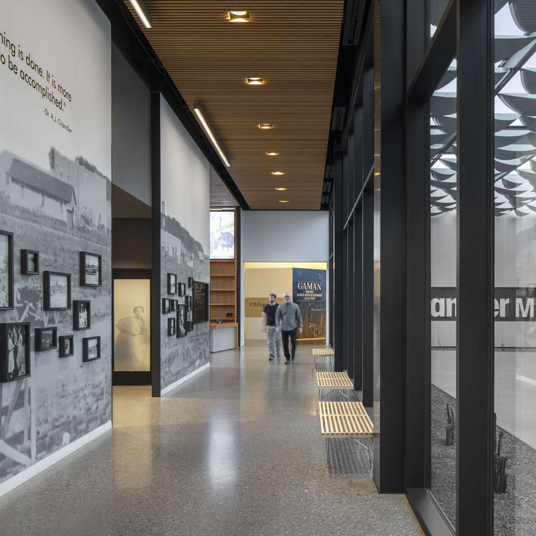 A modern museum corridor with photo displays on the left wall, two people walking in the distance, and large windows on the right filling the space with natural light.