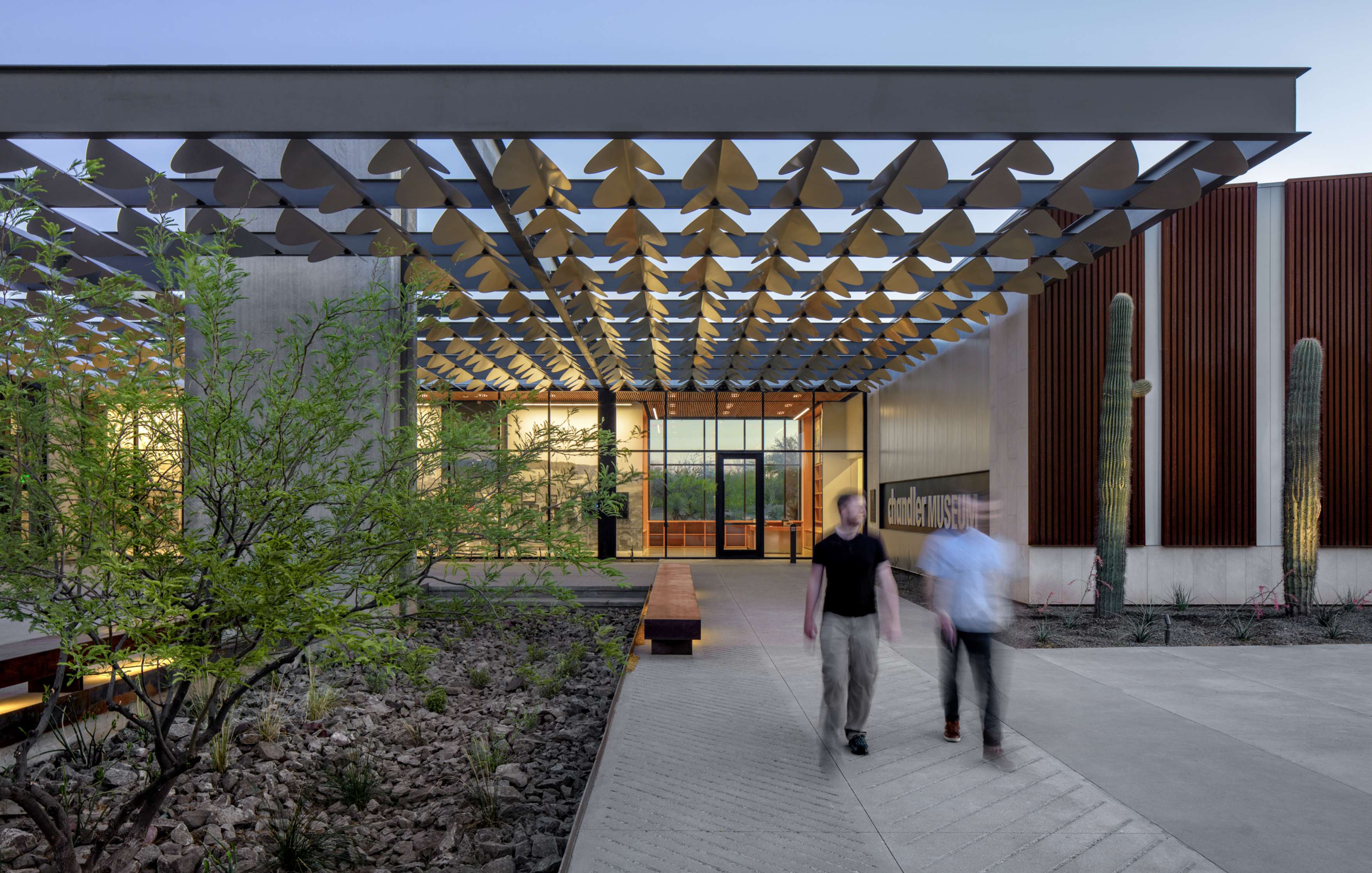 Two people walk on a path near a modern building entrance with a lattice-style canopy, desert plants, and cacti under a clear blue sky.