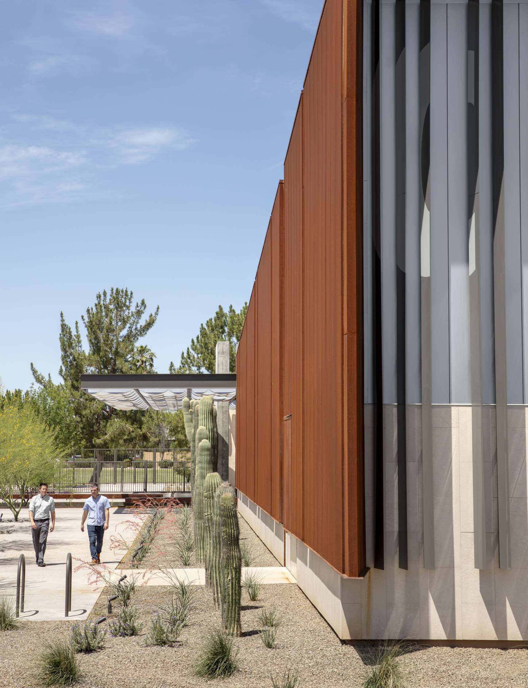 Two people walk along a pathway beside a modern building with rust-colored panels and vertical gray fins. Surrounding the pathway are desert plants and cacti. The sky is clear and blue.