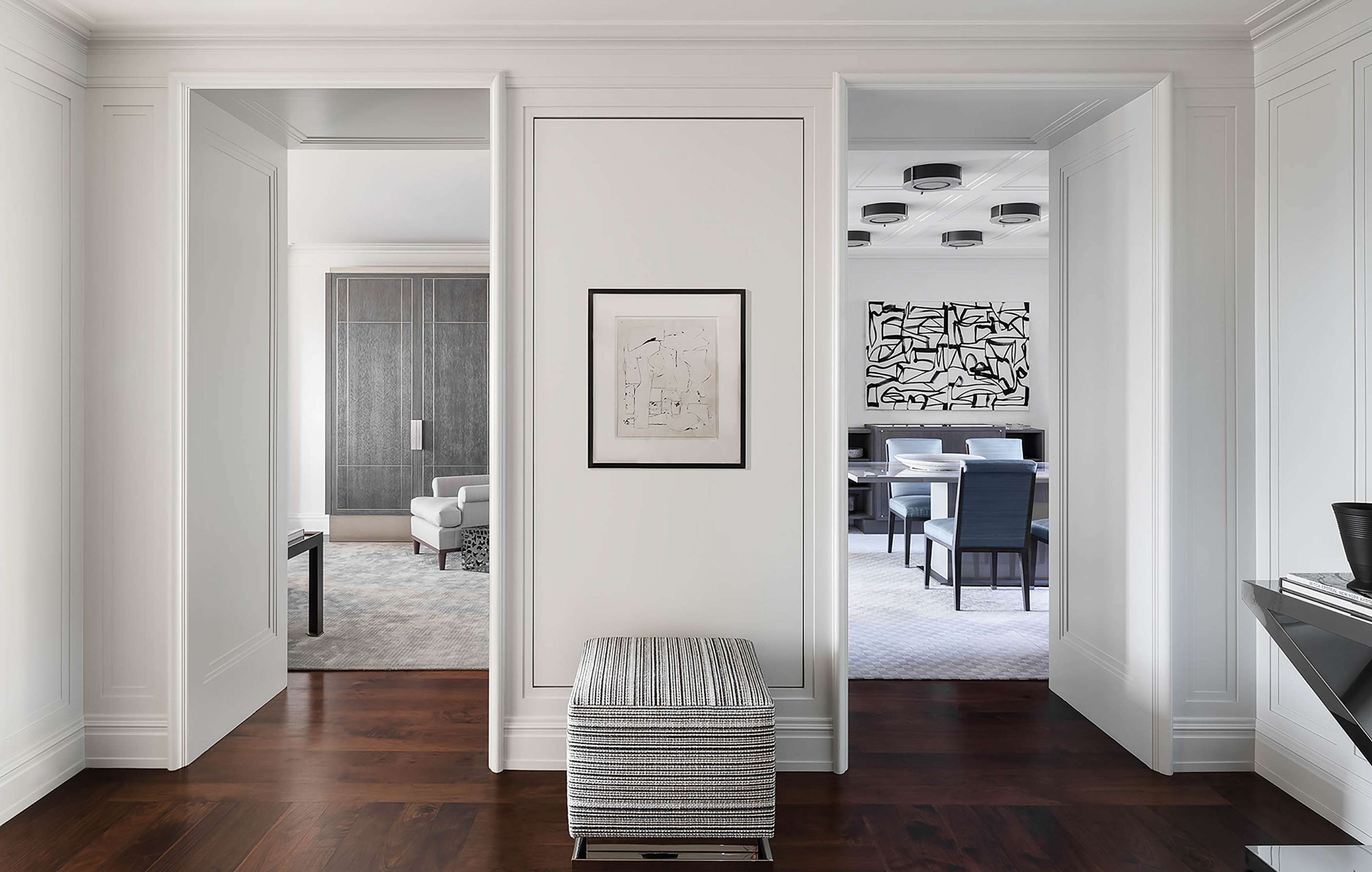 Modern interior with white walls, dark wood flooring, and abstract art. Two open doorways lead to living areas with contemporary furniture and decor. A striped ottoman sits by the wall.