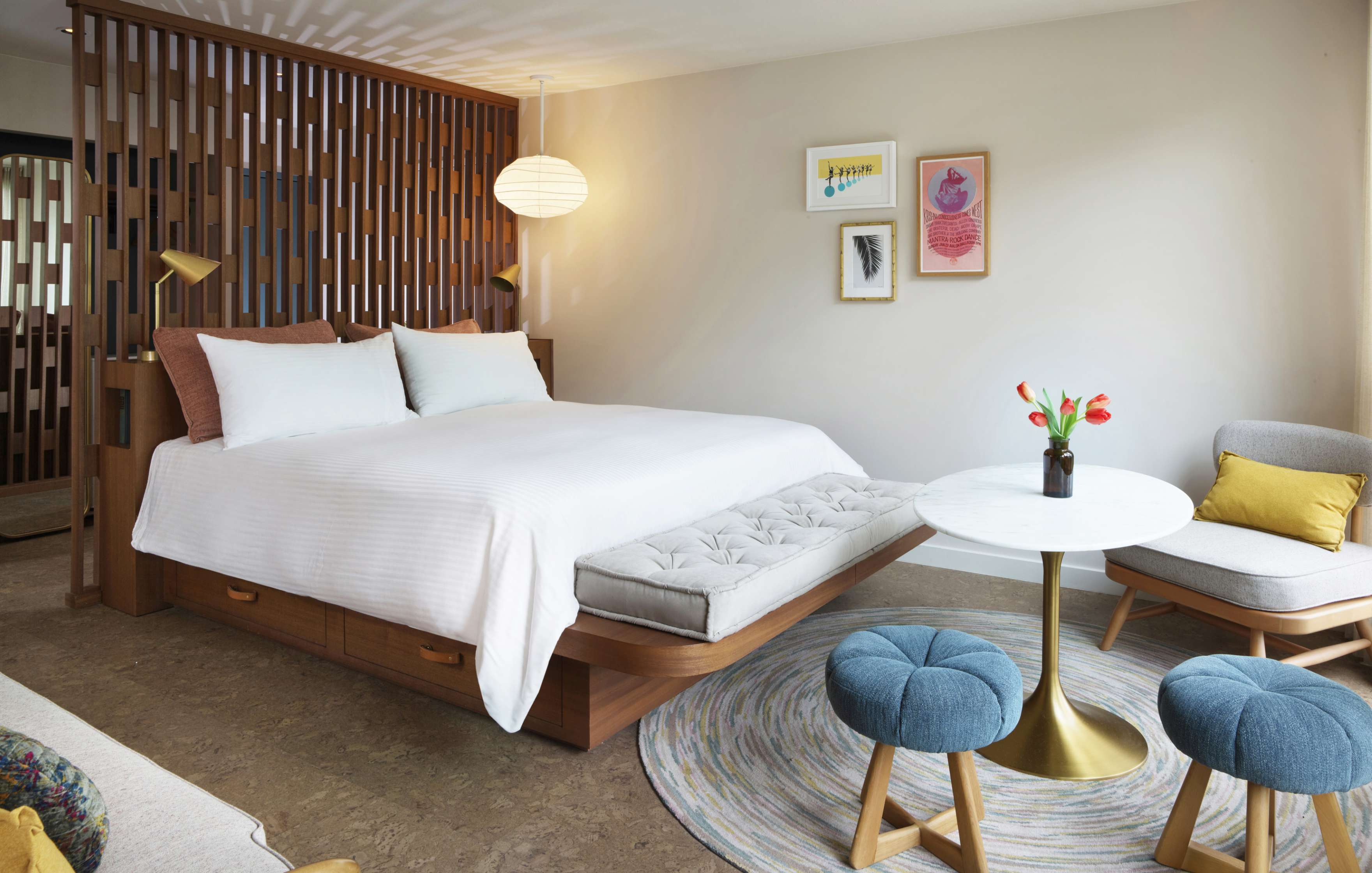 A modern bedroom with a large bed, a wooden partition, wall art, a round table with chairs, and a vase with flowers. Soft neutral tones and natural light create a cozy ambiance.