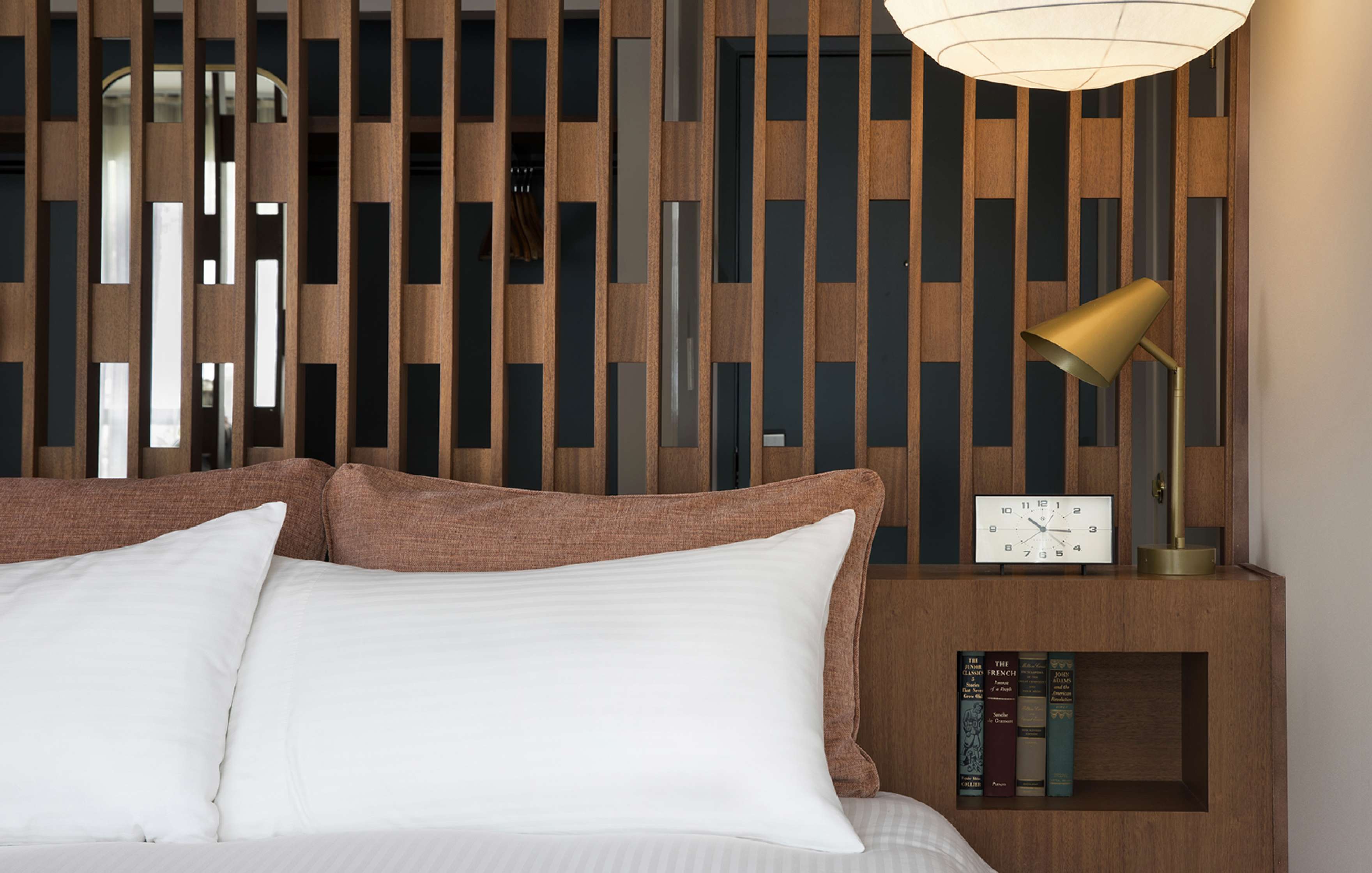 A modern bedroom with a wood slat partition behind a bed, white pillows, a small clock, books, and a gold reading lamp on a built-in shelf.