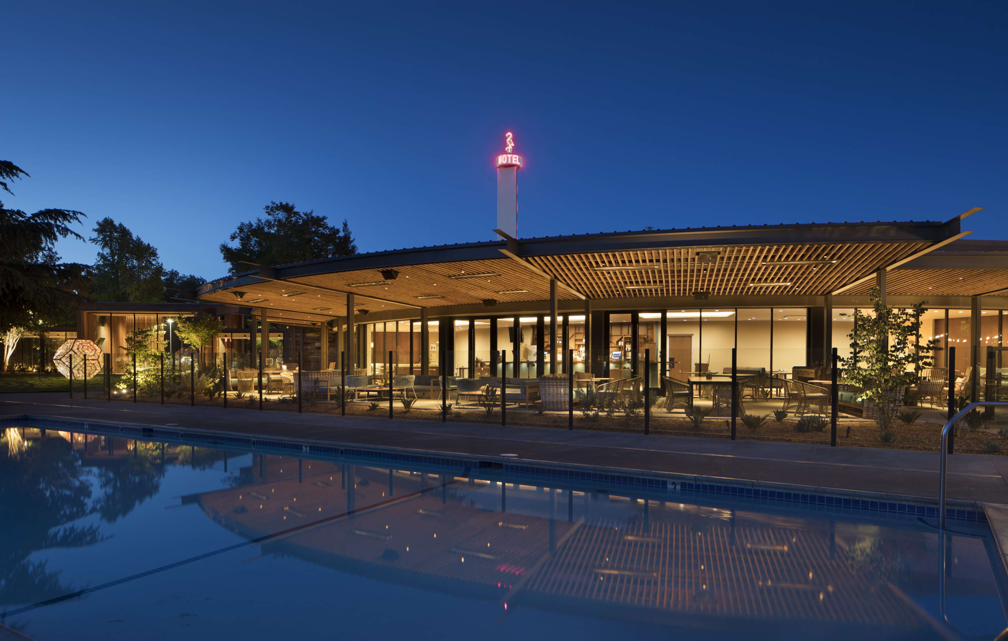 Modern building with large glass windows and wooden pergola, illuminated at dusk, reflecting on a clear pool in the foreground.